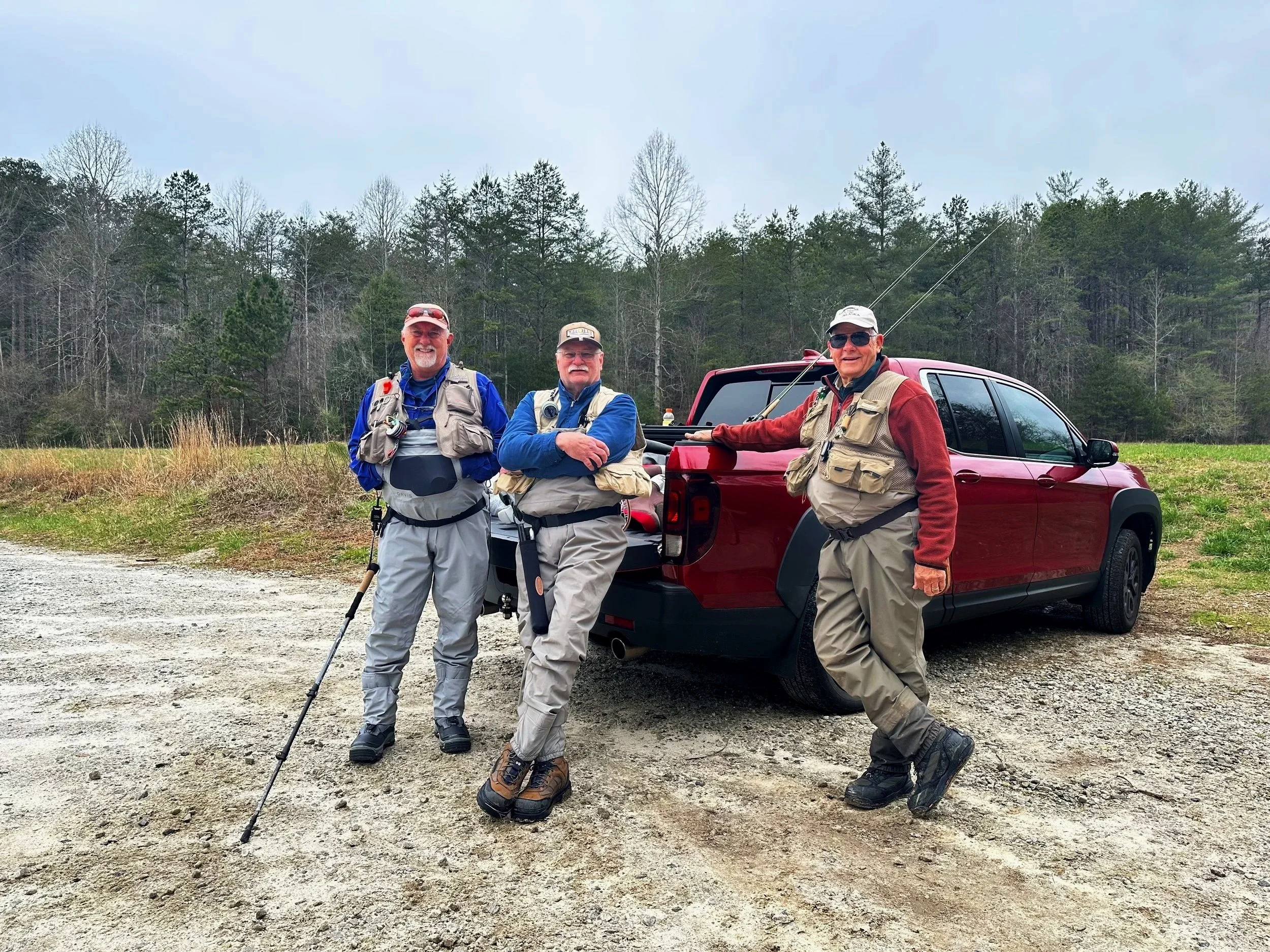 Three men standing next to a red pickup truck in a rural outdoor setting, dressed in outdoor clothing and gear, holding fishing poles.