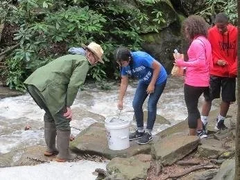 Four people collecting water from a river using a bucket, in an outdoor setting with rocks and greenery.