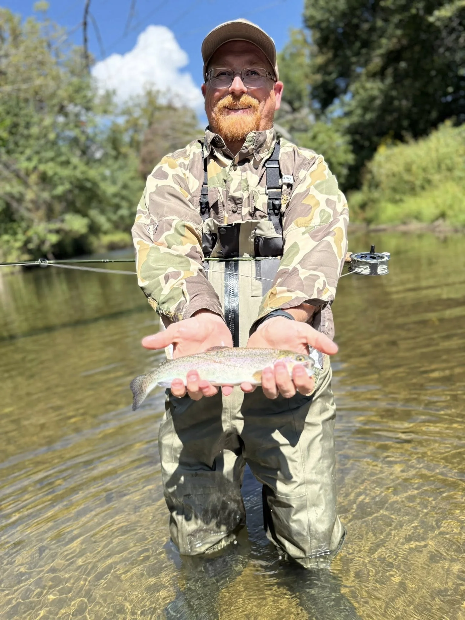 A man standing in a shallow river holding a fish with both hands. He is wearing camouflage outdoor clothing, glasses, a cap, and smiling. There are trees and a partly cloudy sky in the background.