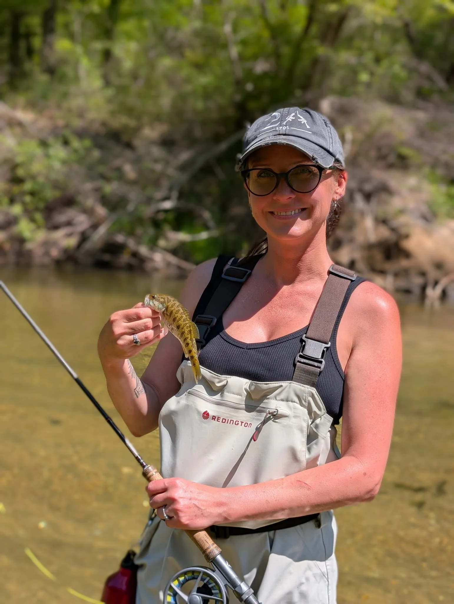 Woman smiling while holding a fish she caught while fishing in a river with green trees in the background.