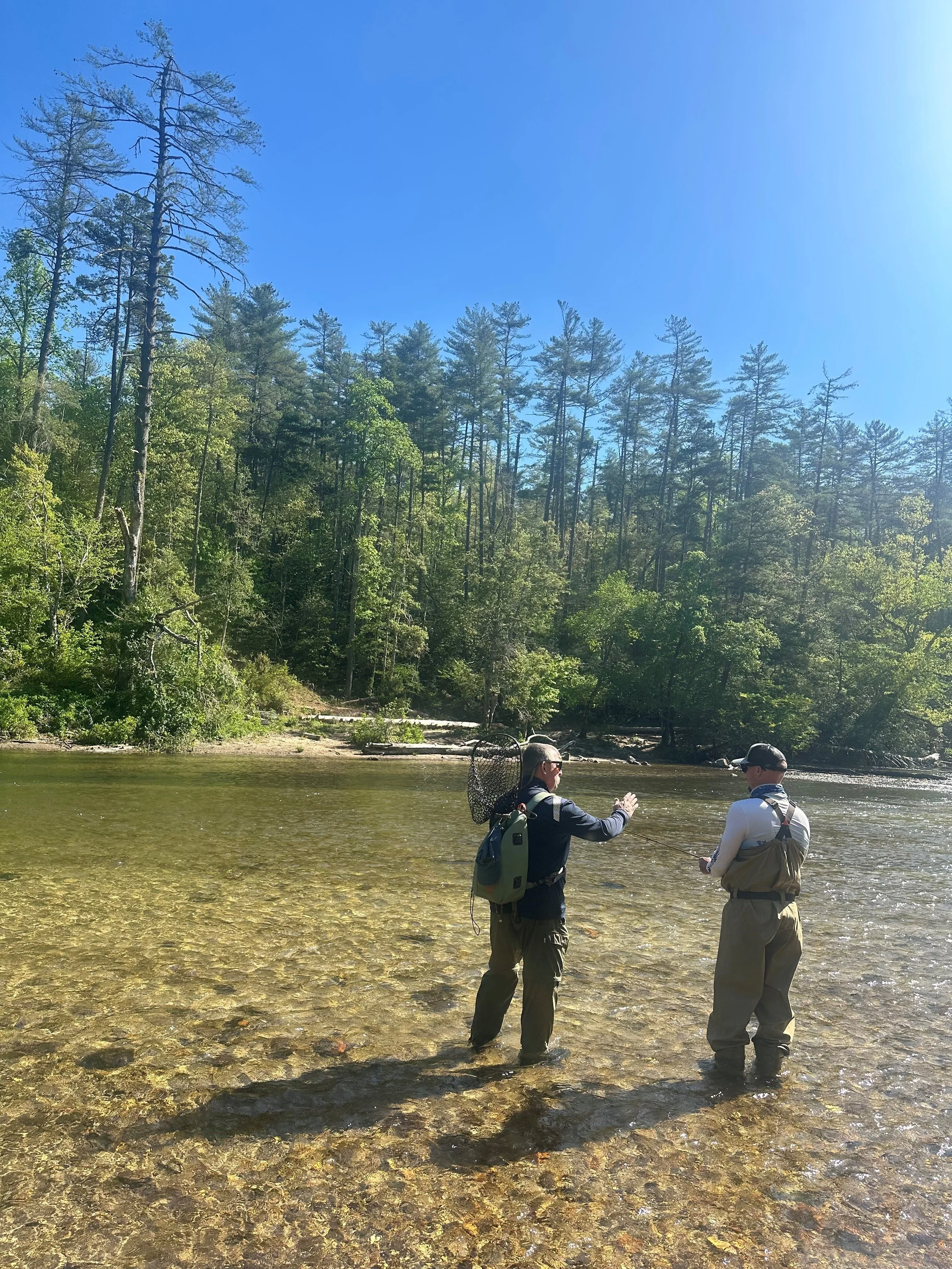 Two people are standing in a shallow river, fishing, with a background of green trees and a clear blue sky.