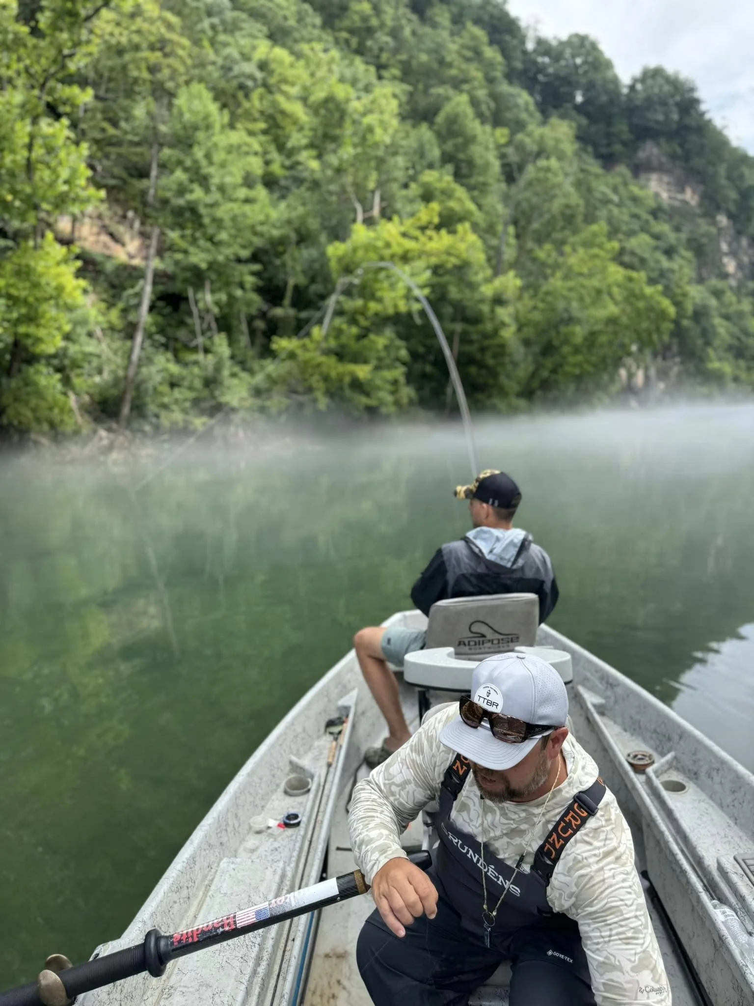 Two men fishing from a boat on a misty river with lush green trees on the riverbank.