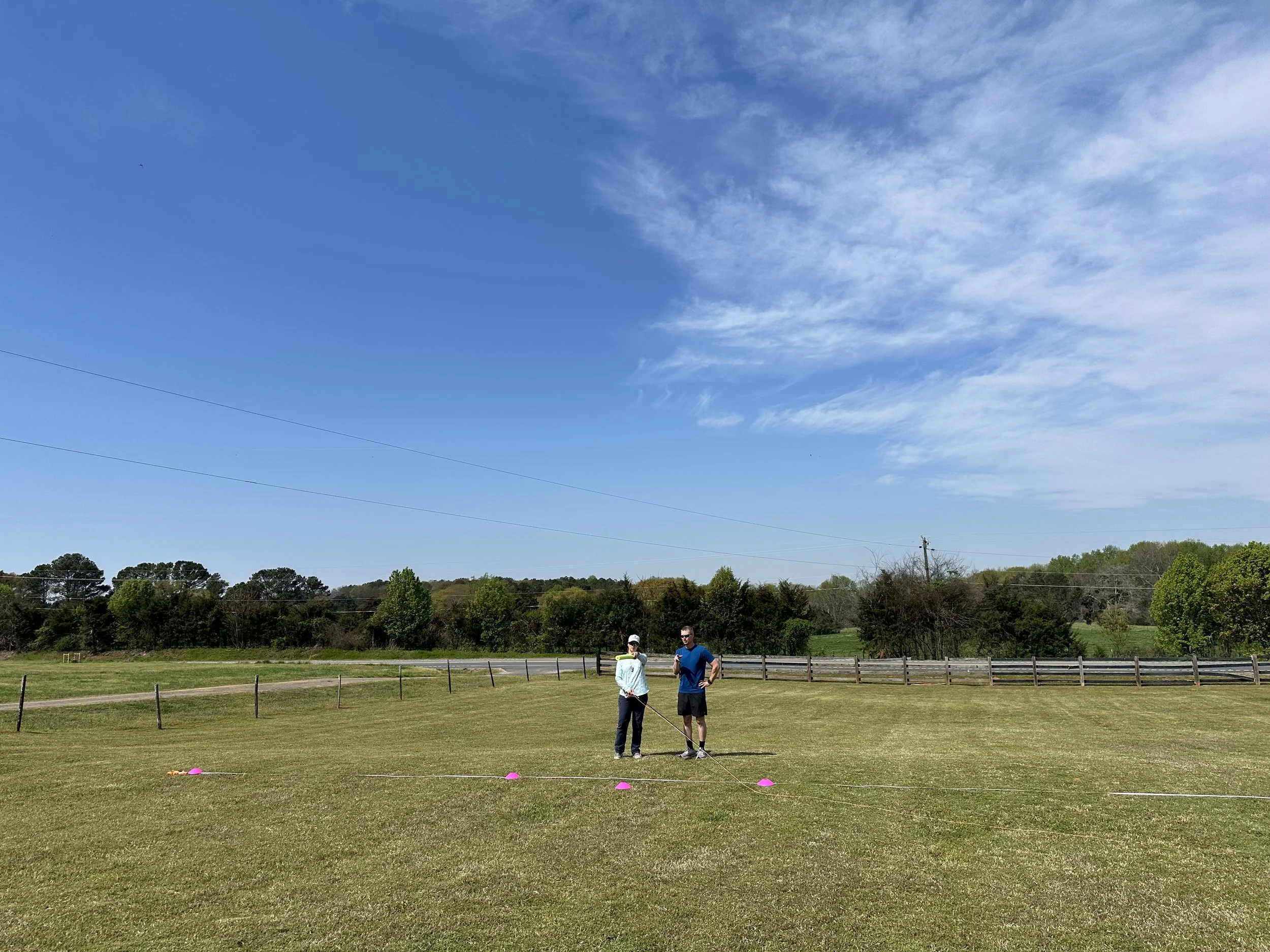 Two people standing on a large grassy field near pink cones, with a blue sky and trees in the background.