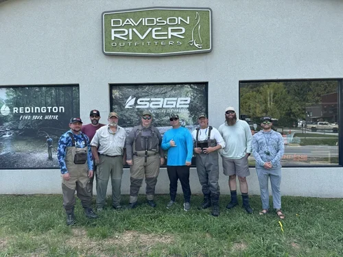 Group of eight men standing outside in front of a building with signs for Davidson River Outfitters, Sage, and Redington.