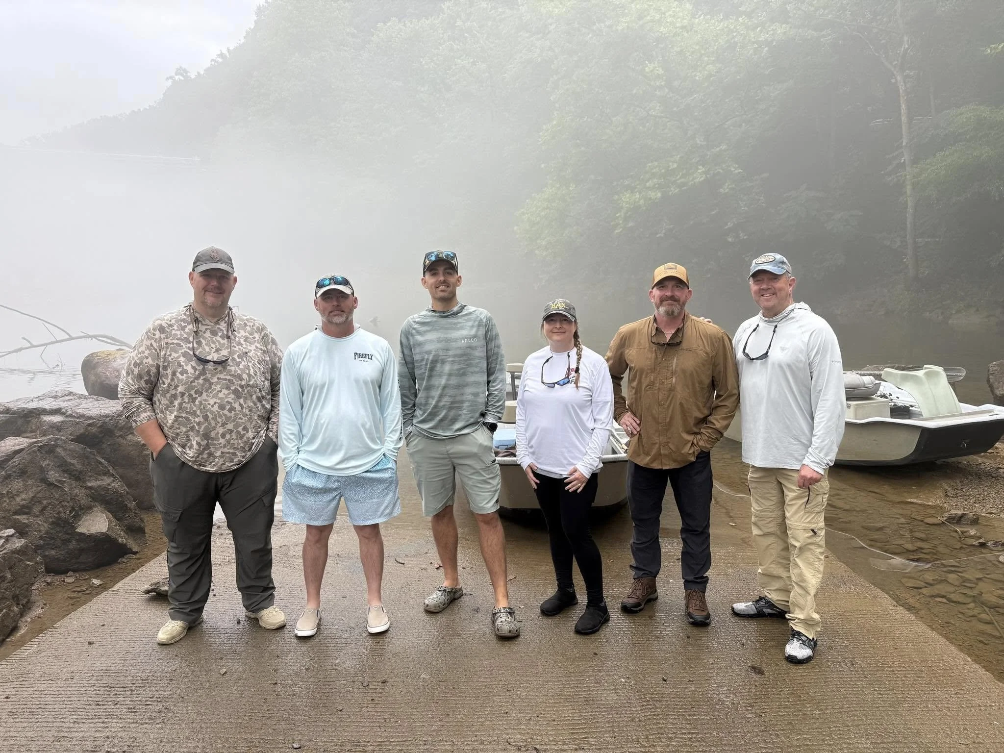 Six people standing on a concrete pier with boats and foggy water in the background, outdoors in a natural setting with trees.