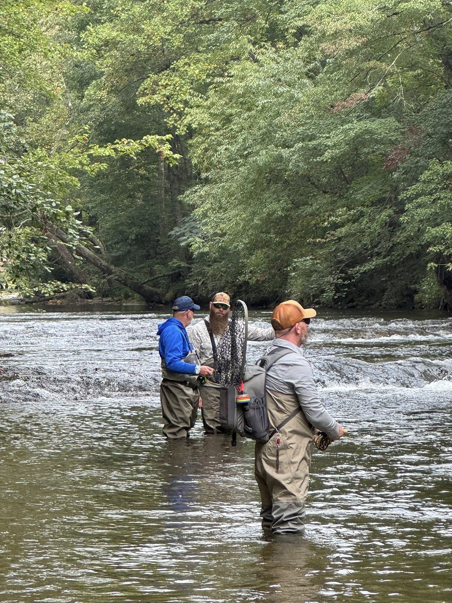 Three men fishing in a river surrounded by trees, with one man holding a fishing rod and the other two with fishing gear.