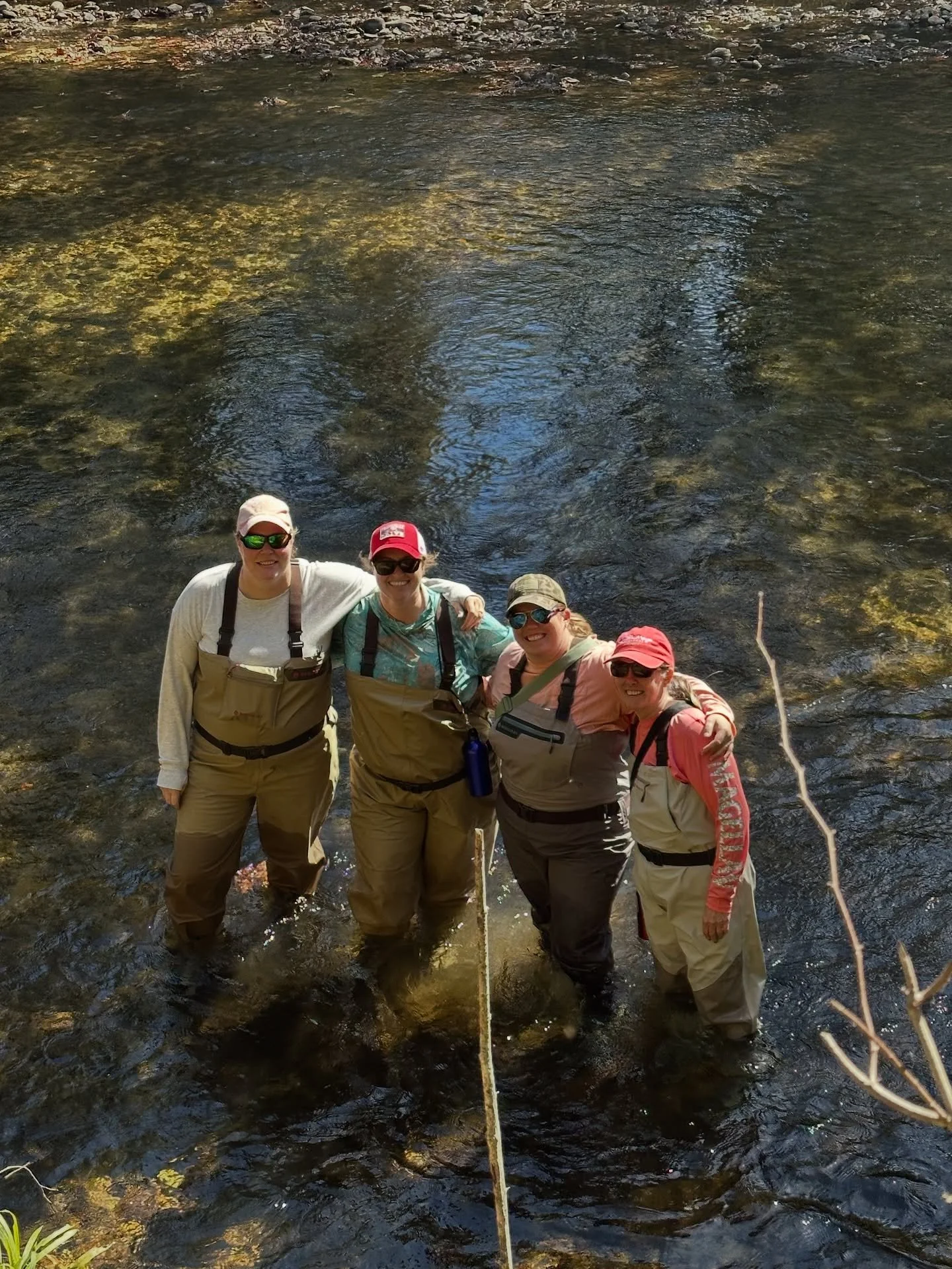 Four women wearing waders and sunglasses standing in a shallow river, smiling and holding onto each other.