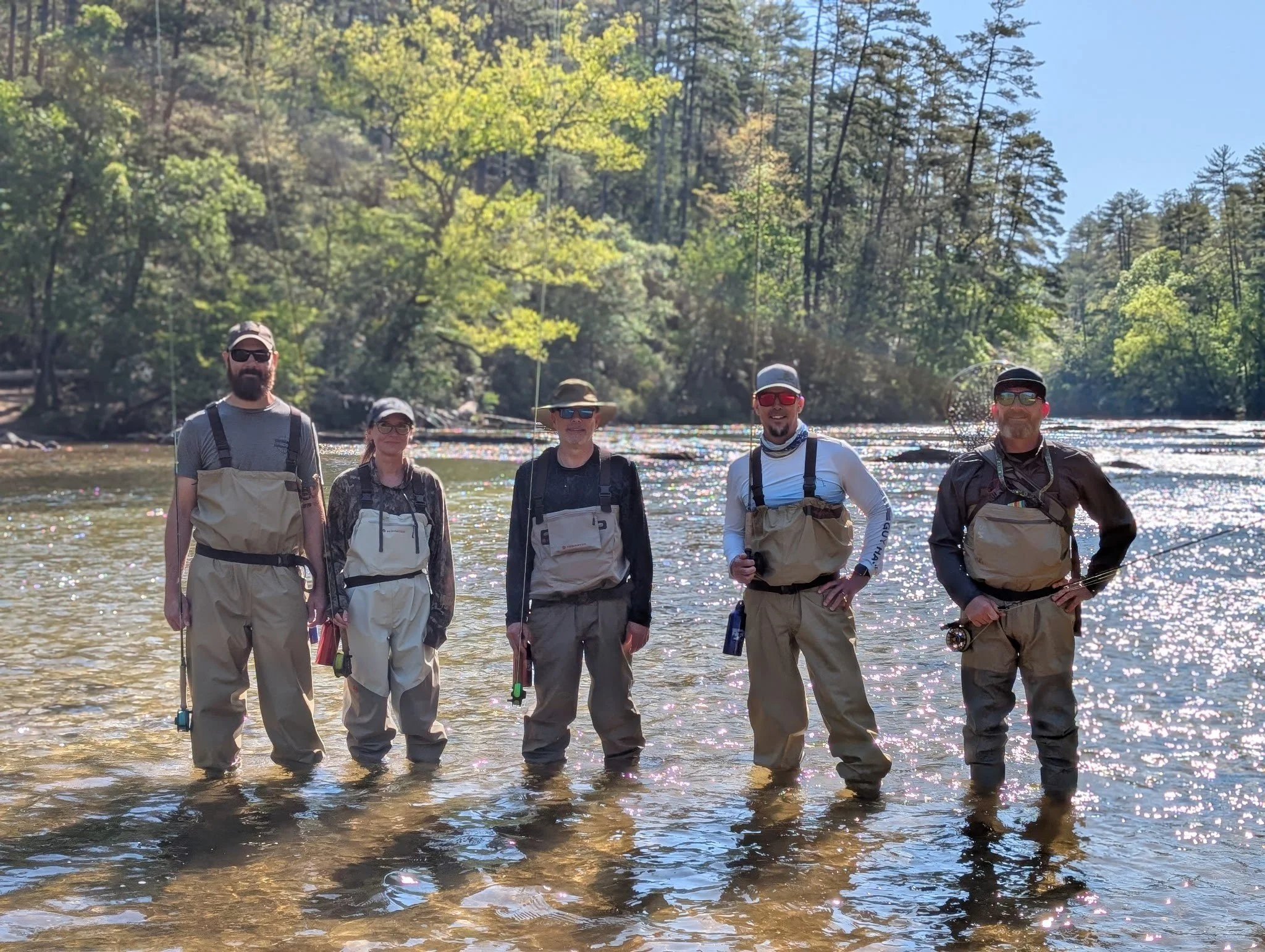 Five people standing in shallow water near a river, smiling and wearing fishing gear. There are trees with green leaves and a clear blue sky in the background.