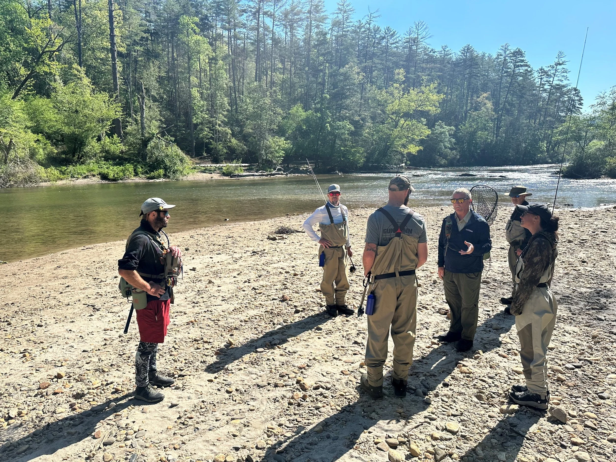 A group of six people standing on a rocky beach by a river, surrounded by trees, engaged in conversation, some holding fishing rods, on a sunny day with clear blue skies.