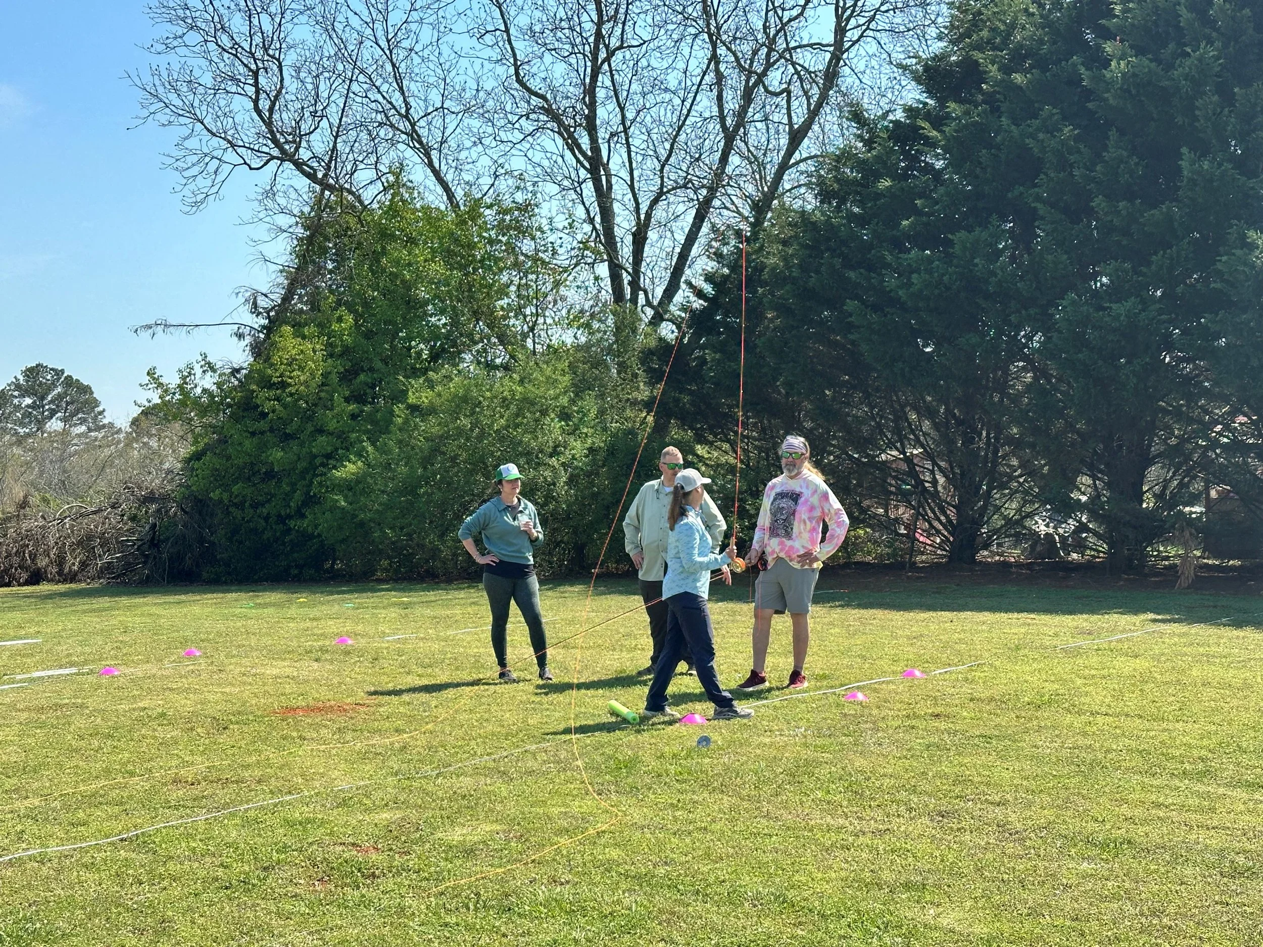 Four people standing on a grassy field with trees in the background, participating in an outdoor activity that involves measuring or setting up with strings and cones.