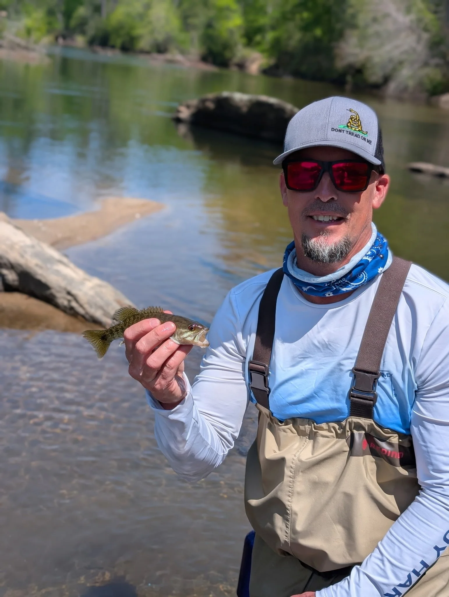 A man wearing sunglasses and a cap holding a fish in front of a river with rocks and trees in the background.