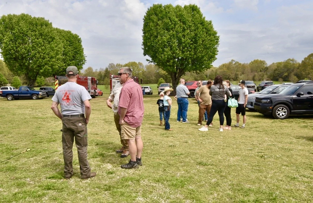 People gathered outdoors on a grassy field near parked cars, with trees in the background, on a partly cloudy day.