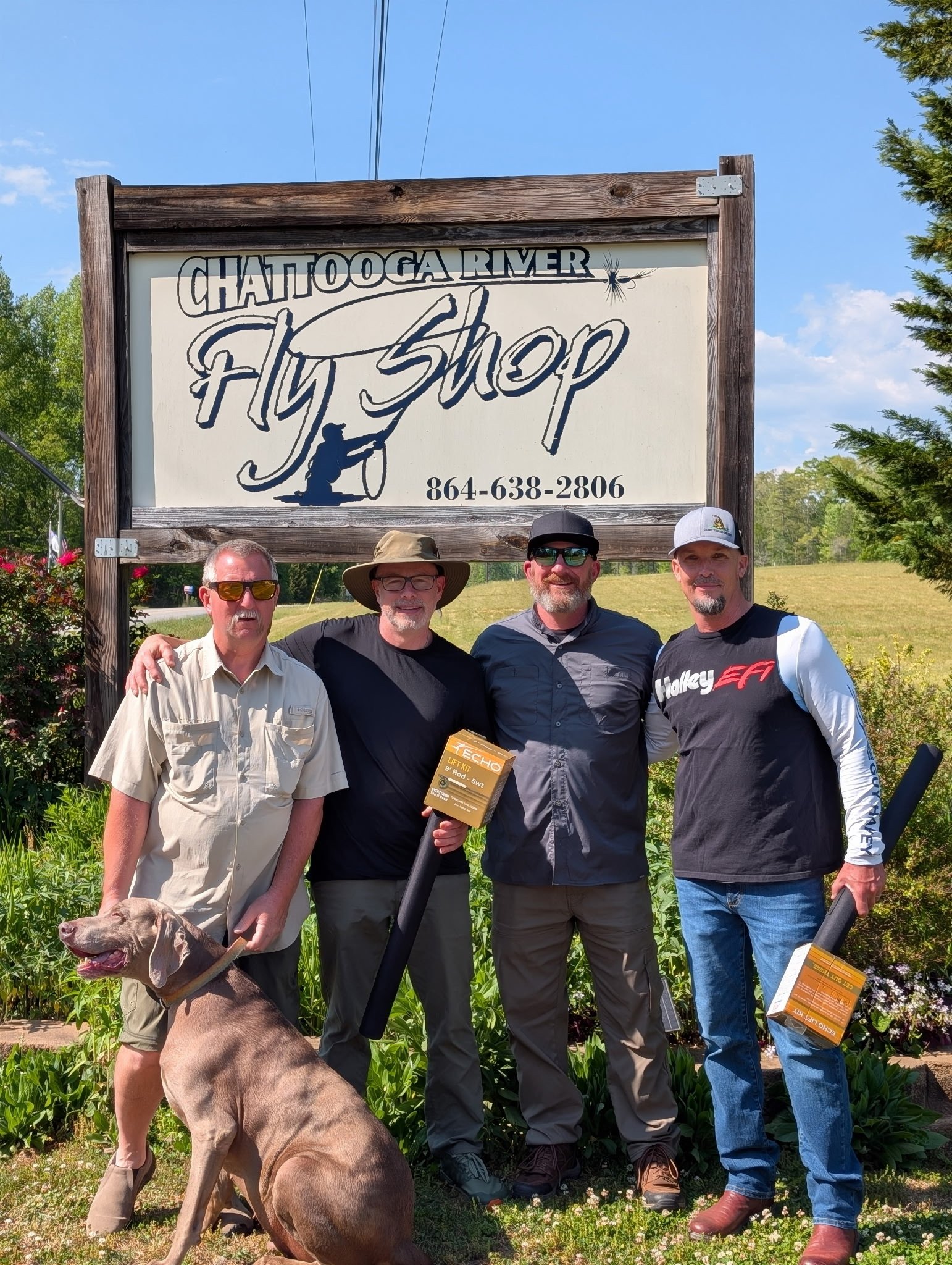 A group of four men standing outdoors in front of a sign that reads 'Chattanooga River Fly Shop' with a phone number underneath. The men are smiling and posing for the photo, with one holding a box labeled 'Echo Lift Kit.' A dog is in the foreground,