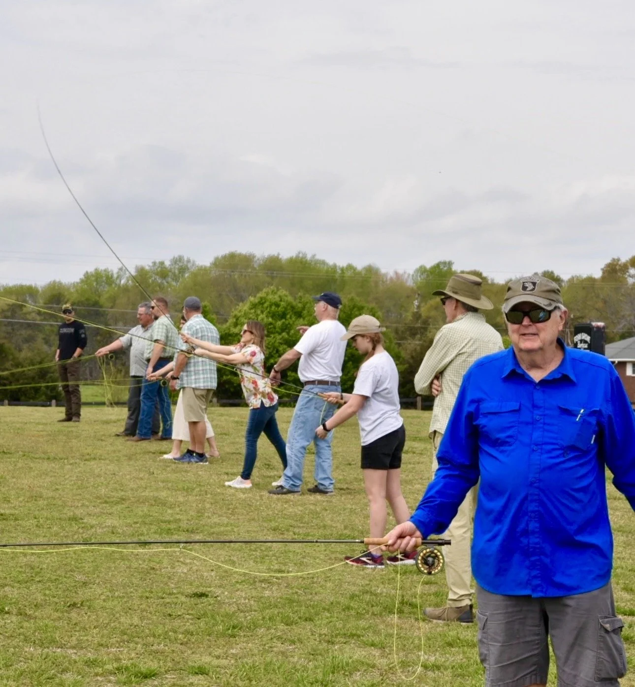 Multiple people standing outdoors in a line, holding fishing rods during daytime, with a cloudy sky and green trees in the background.