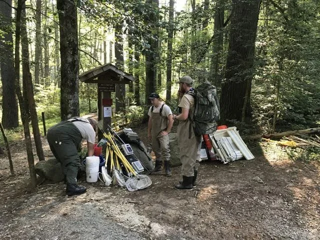 Three people at an outdoor trailhead in a forest, preparing equipment and supplies, with various gear and signs around.