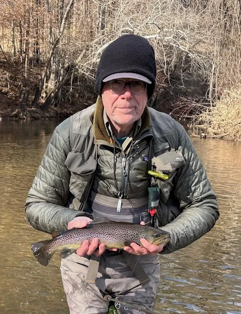 A man dressed in outdoor gear, including a beanie, glasses, and a life jacket, stands in a river holding a large fish he caught, with a forested area in the background.