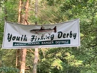 A banner hanging between trees with a shark graphic and the text 'Youth Fishing Derby' and 'Andrew Pickens Ranger District'.