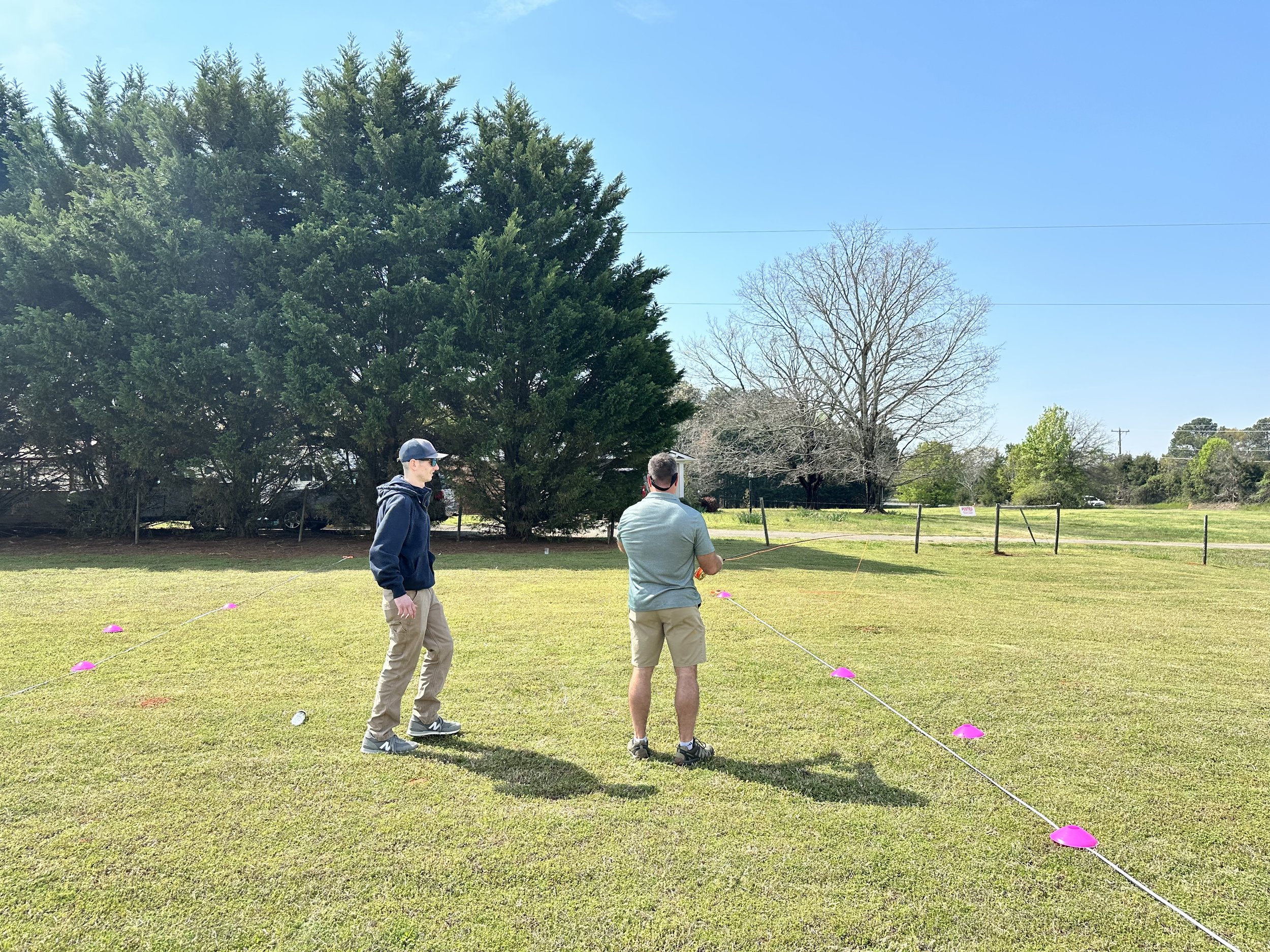Two men stand on a grassy field near a line of pink cones, with trees and a blue sky in the background, during a sunny day.