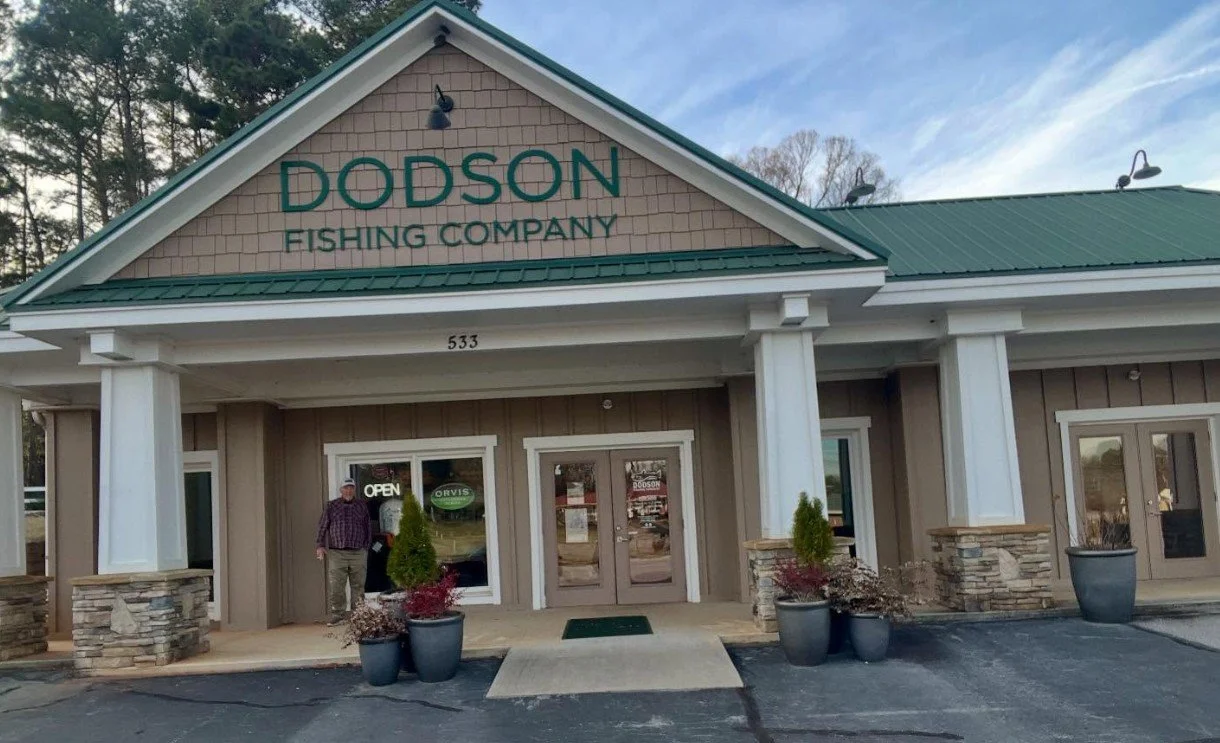 Exterior of Dodson Fishing Company store with a green metal roof and beige siding, two large potted plants, and a person standing near the window.