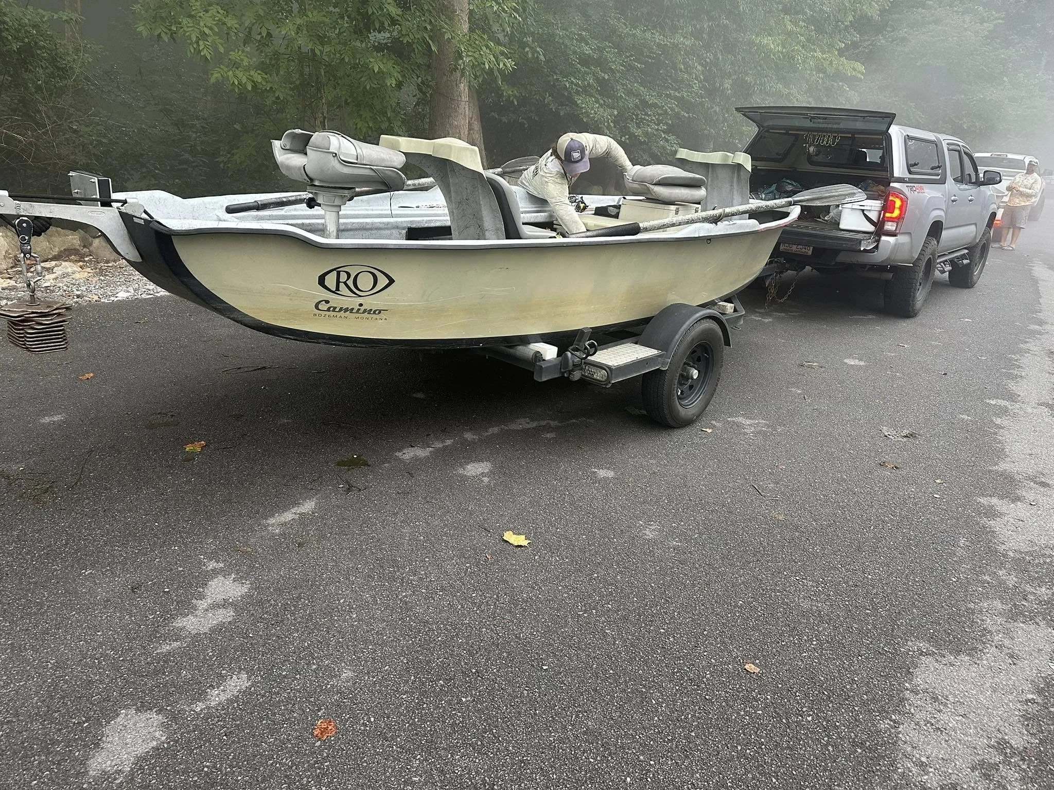 A pickup truck with an open tailgate towing a small boat on a trailer. A man is working inside the boat, and another person stands in the background near the truck. The scene is outdoors on a paved road with trees overhead.