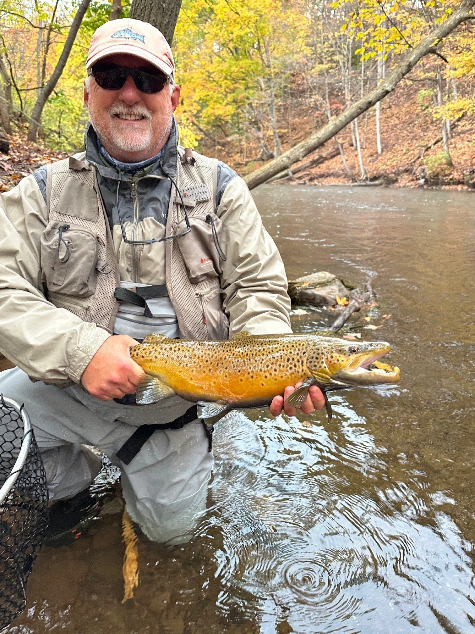 A man in outdoor fishing gear holding a large trout fish by a river surrounded by fall-colored trees.