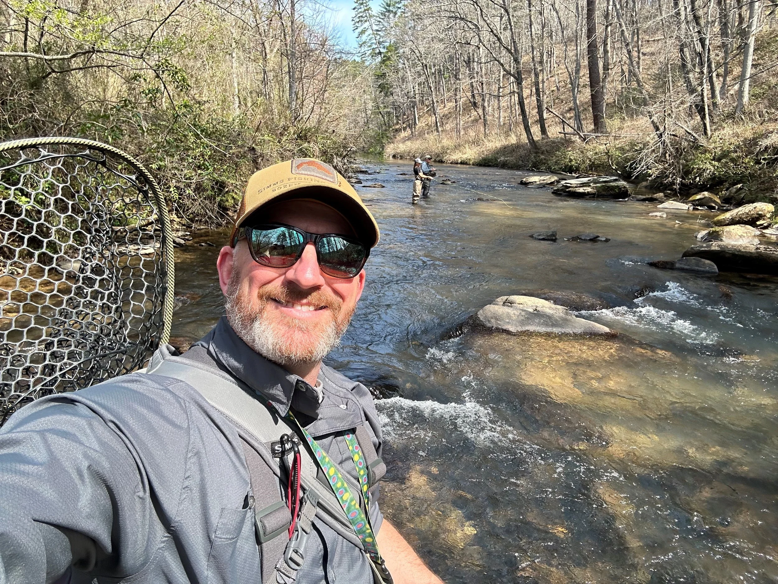 A man taking a selfie during a fishing trip by a river, with two other fishermen in the background, surrounded by trees and rocks.