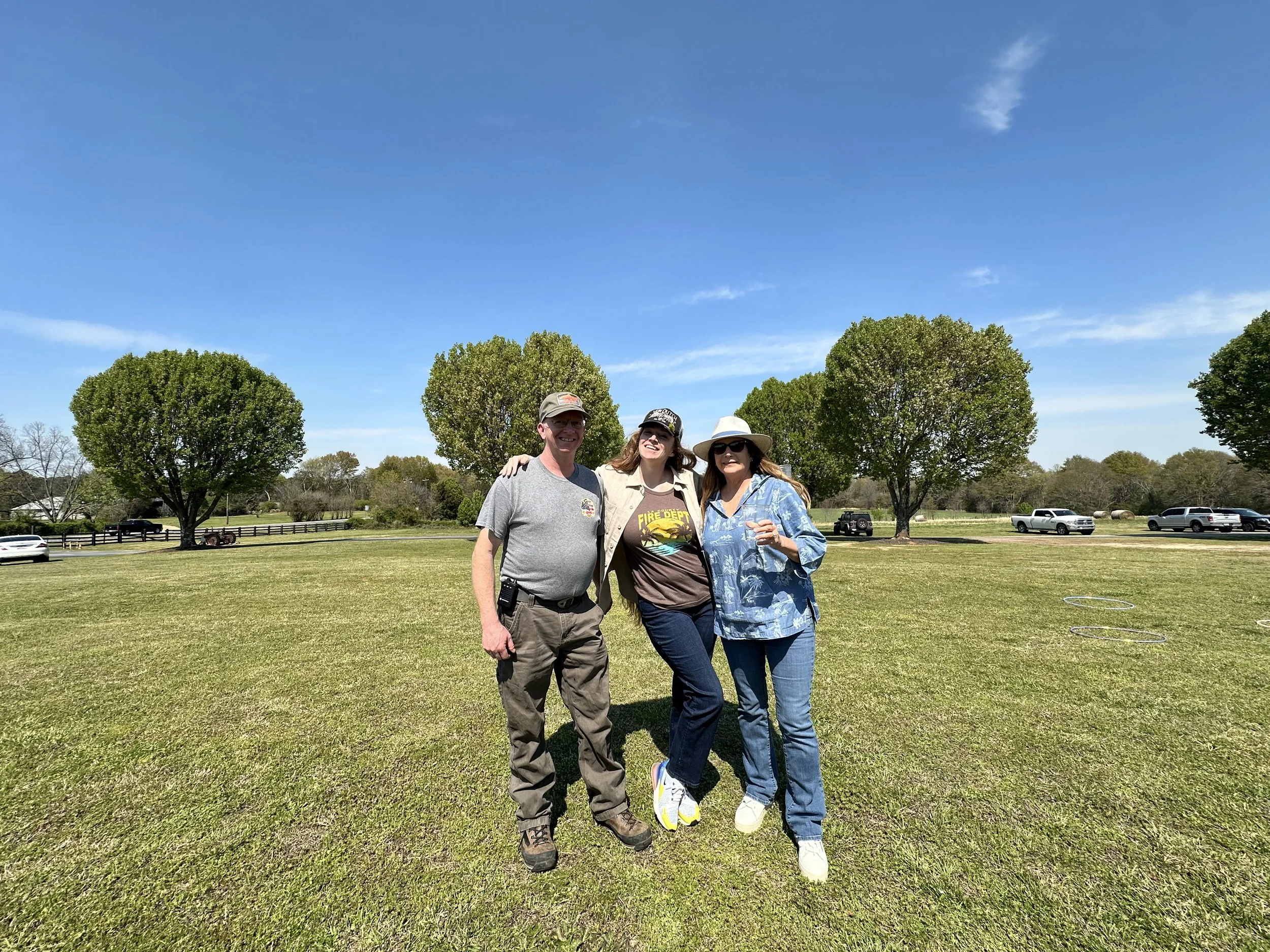 Three people standing outdoors on a sunny day with green grass and trees in the background. The man on the left is wearing a gray t-shirt and khaki pants. The woman in the middle is wearing a tan jacket and dark pants. The woman on the right is weari