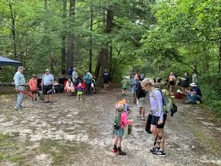 Group of people, including children, gathered outdoors in a wooded area with trees and a wet ground, participating in an event or gathering.