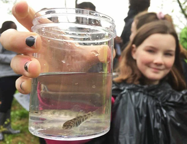 Person holding a clear jar with a small fish inside, with a young girl smiling and standing outdoors in the background.