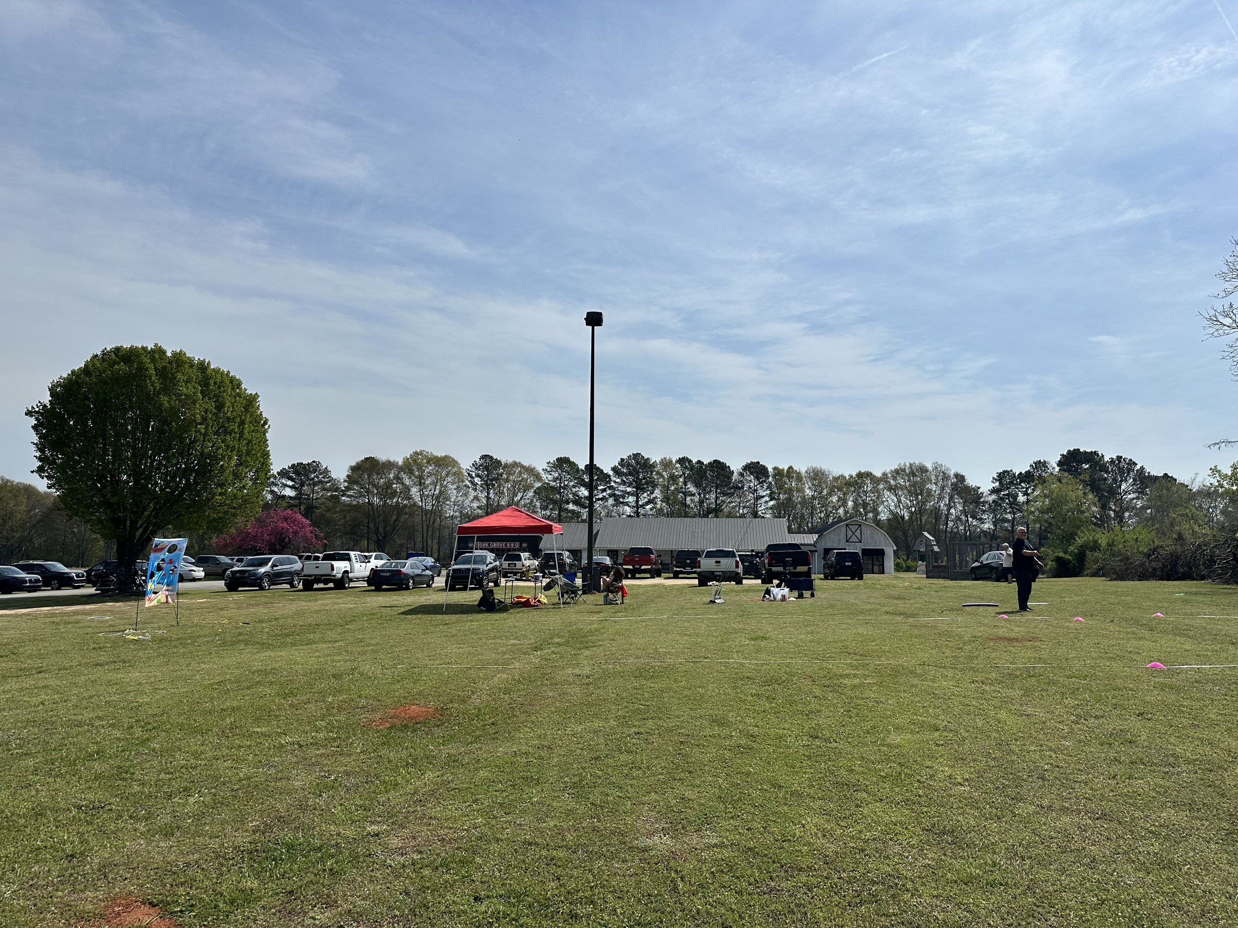Open grassy field with people gathered under a red canopy, parking lot with cars behind, and a small barn structure in the background, under a partly cloudy sky.
