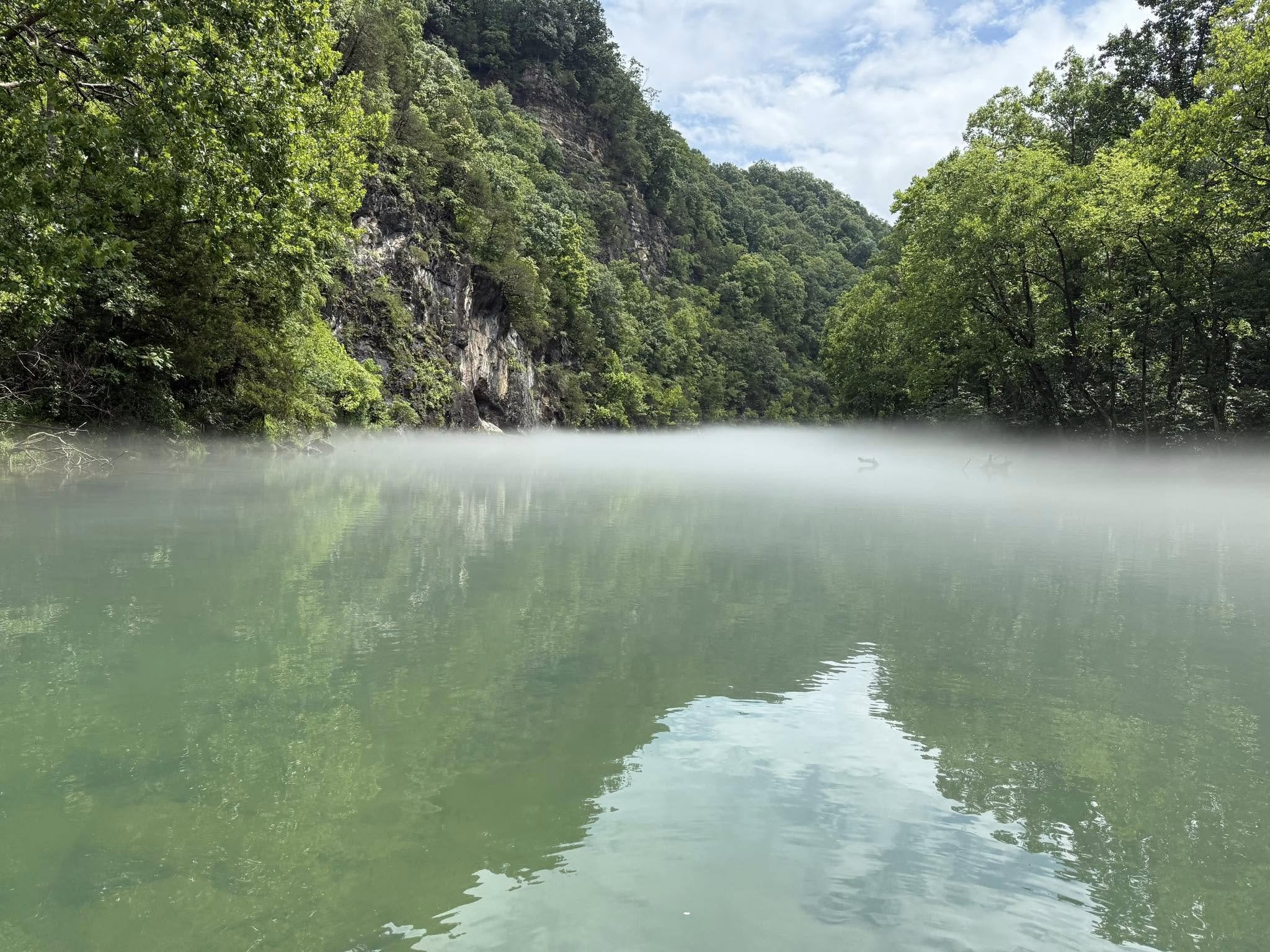 A river with a light layer of fog on the surface, surrounded by green trees and rocky cliffs on the left, with a partly cloudy sky above.