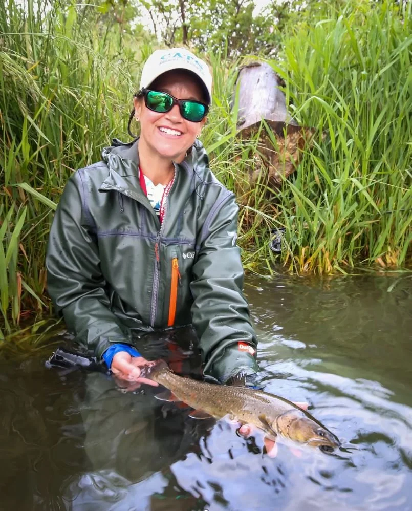 A woman in outdoor gear, sunglasses, and a hat, holding a fish she caught in a river surrounded by tall grass and greenery, smiling at the camera.