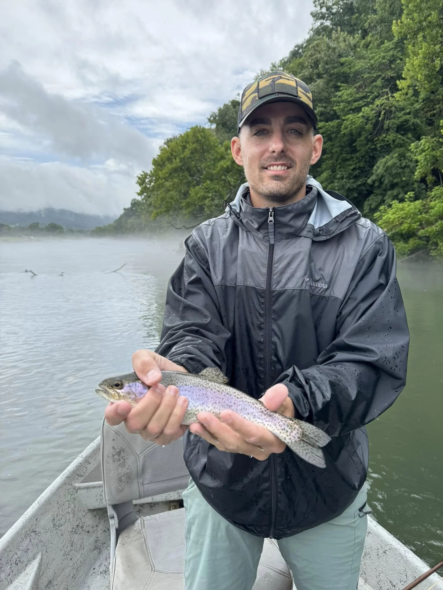 A man in a rain jacket and cap on a boat, holding a rainbow trout he caught, with a foggy river and green trees in the background.