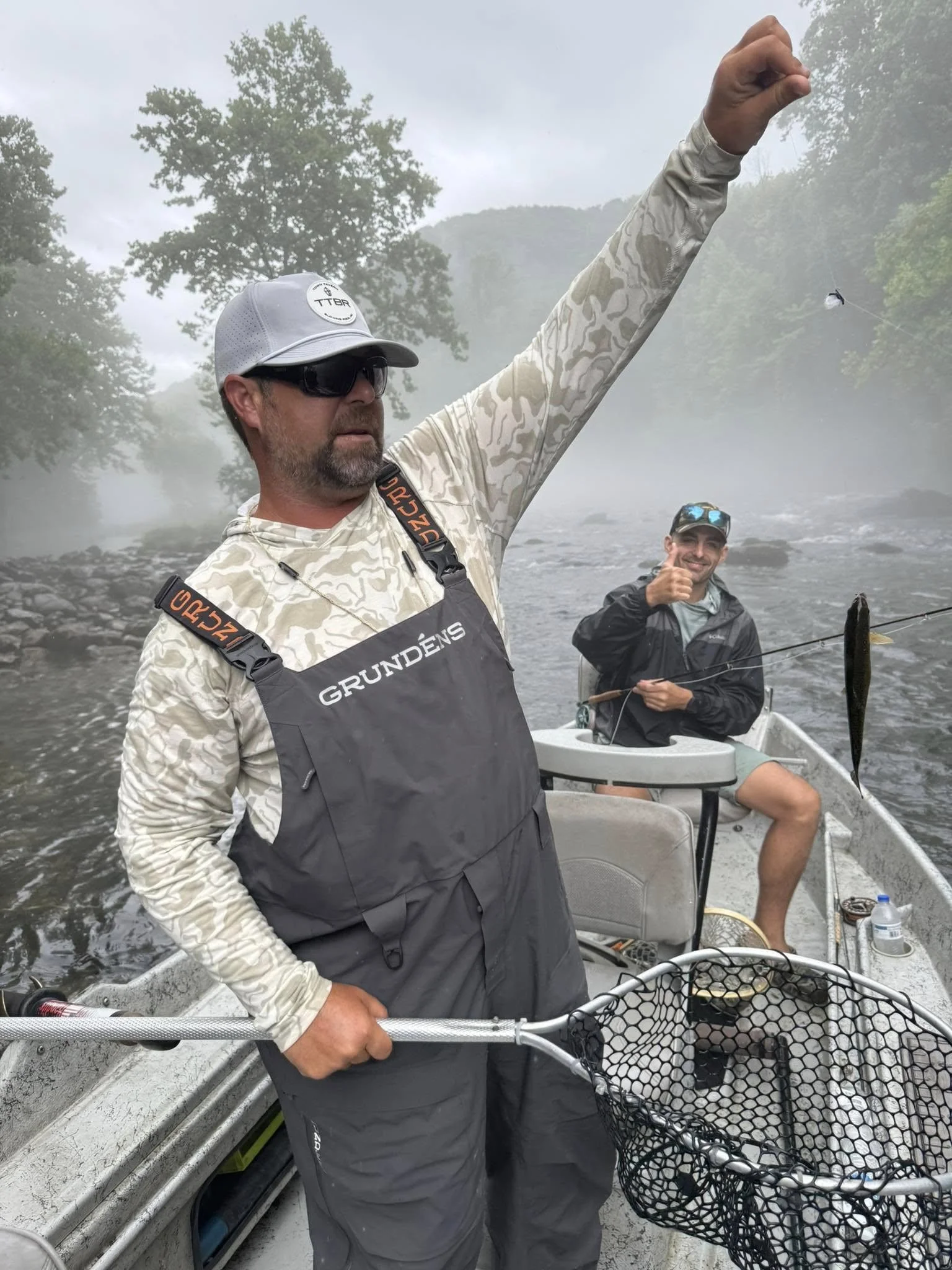 Two men fishing from a boat on a river, with one man in the foreground holding a fishing net and the other in the background giving a thumbs-up. The scene is misty with trees and hills in the background.