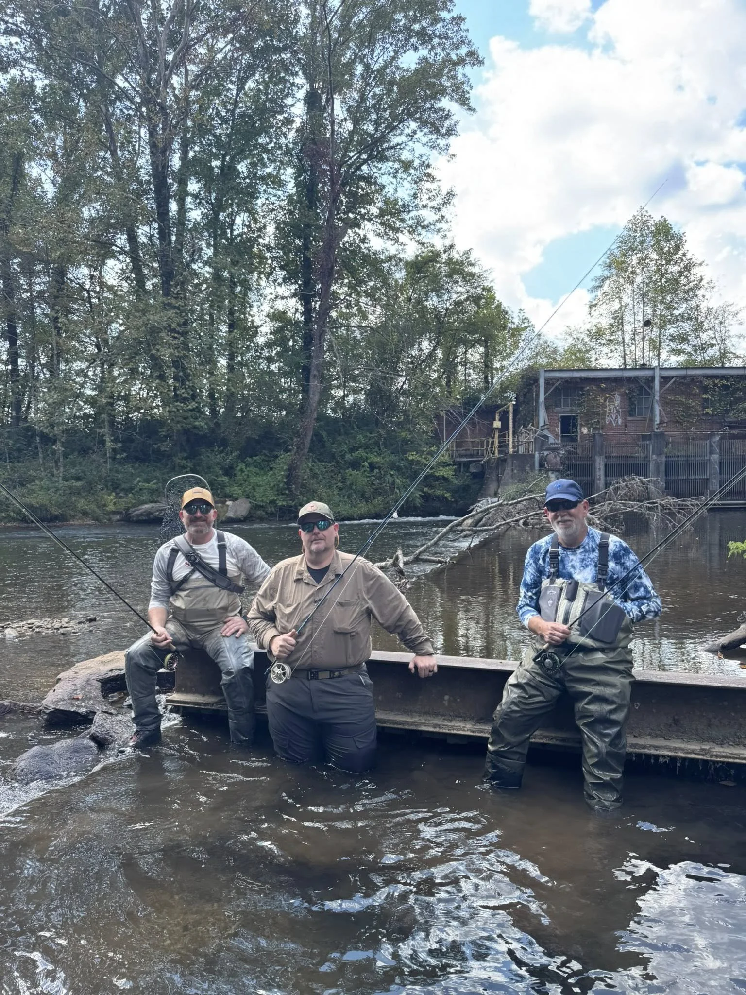 Three men fishing in a river with trees and a building in the background.
