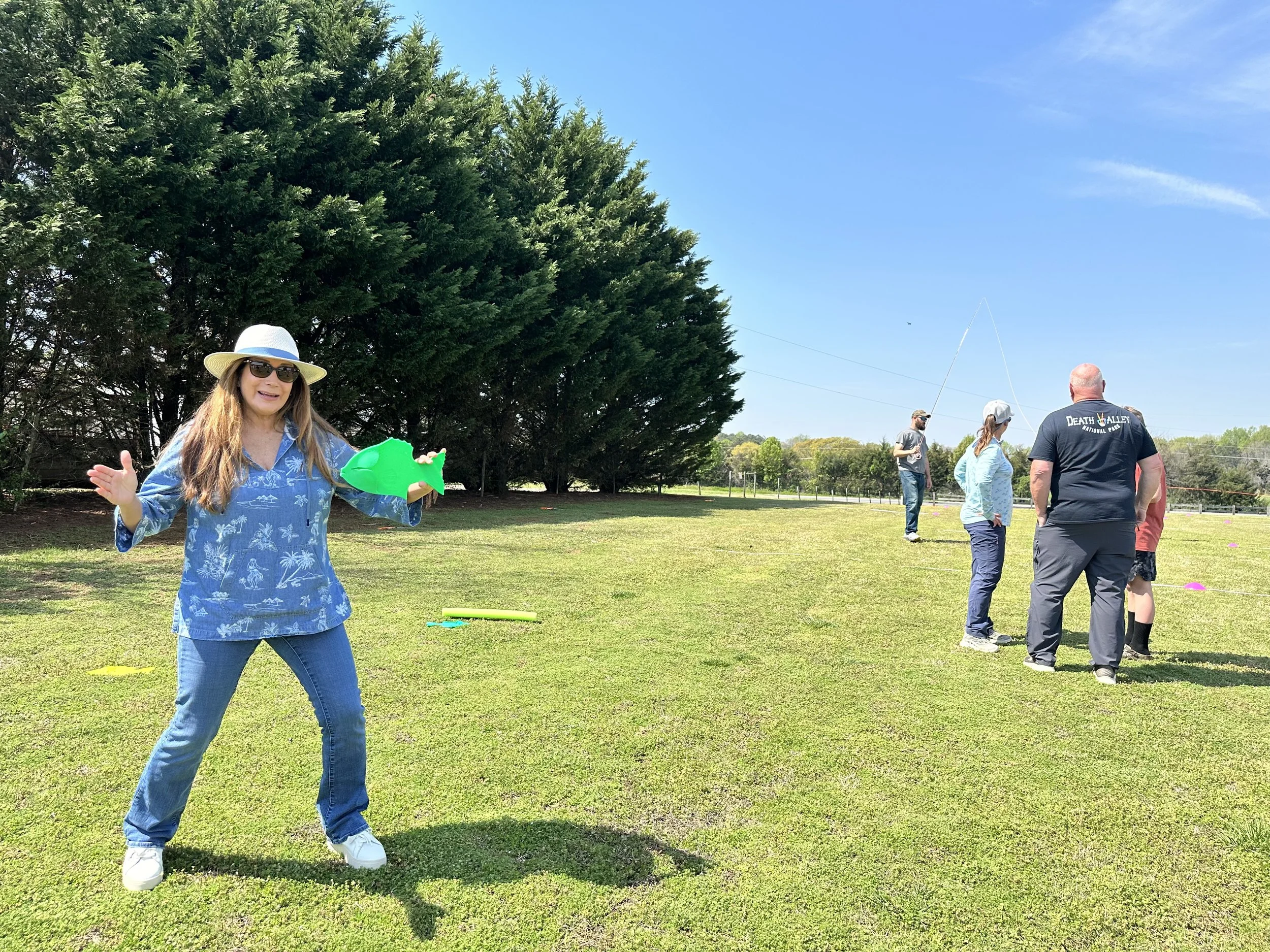 A woman wearing a straw sun hat, sunglasses, a blue tropical-themed shirt, and jeans, holding a green water gun, stands on a grassy field with a group of people fishing in the background under a clear blue sky.
