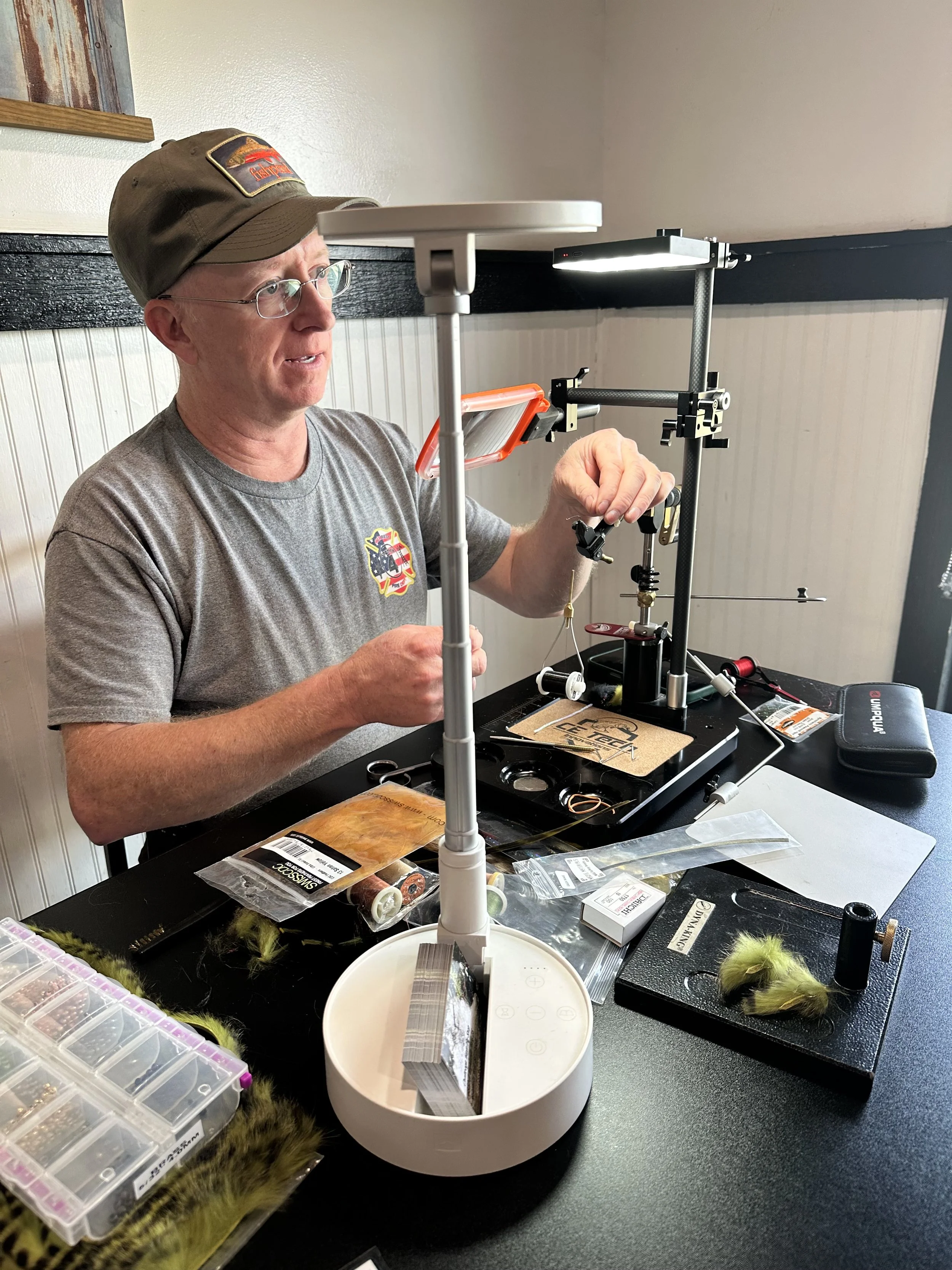 A man wearing glasses, a gray t-shirt with a logo, and a brown cap is sitting at a black table, using fly tying tools. There are various materials, such as feathers and hooks, organized in plastic containers around him as he works on tying a fishing 