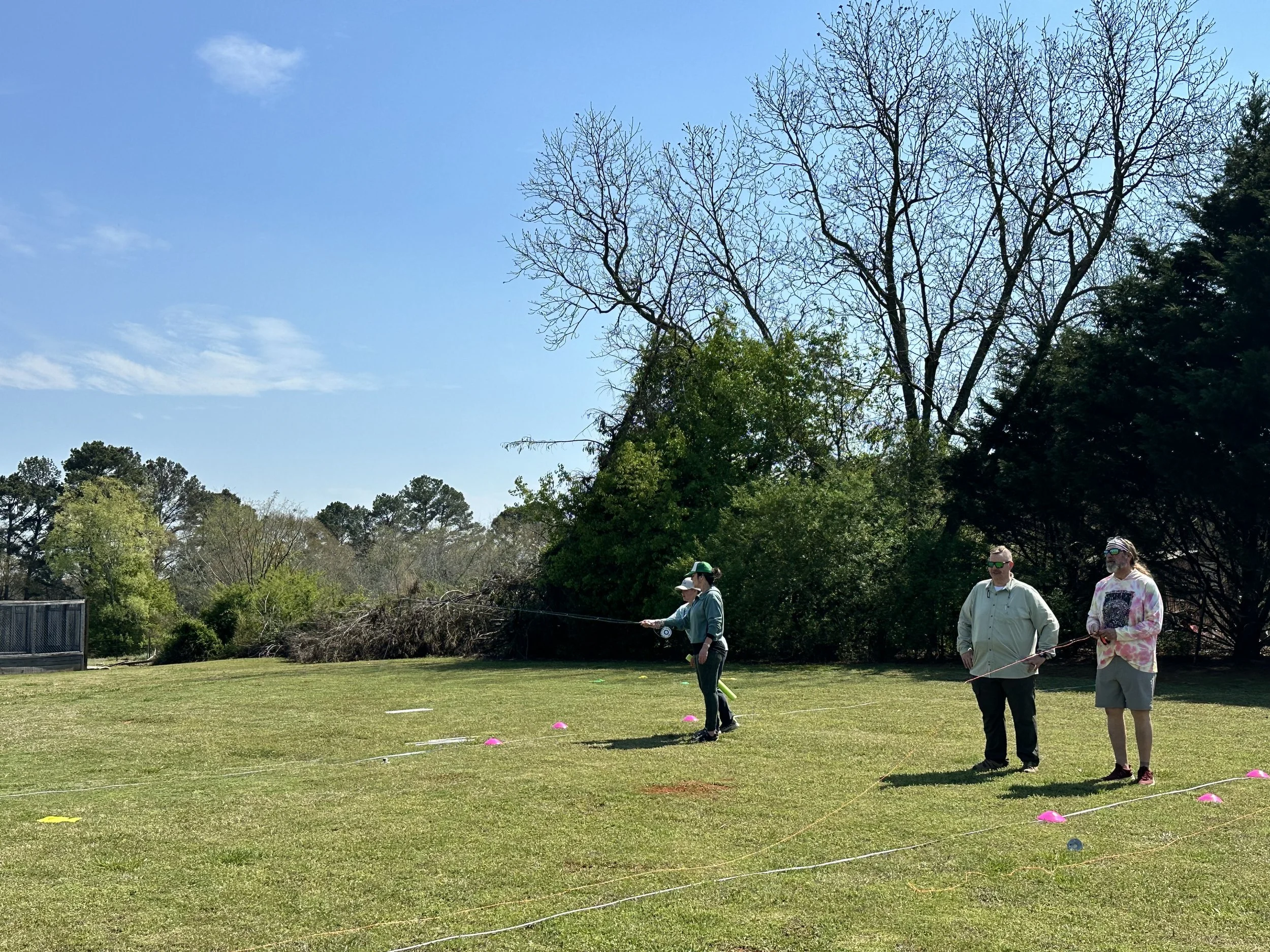Three people standing on a grassy field, holding fishing rods, with trees and a clear blue sky in the background.