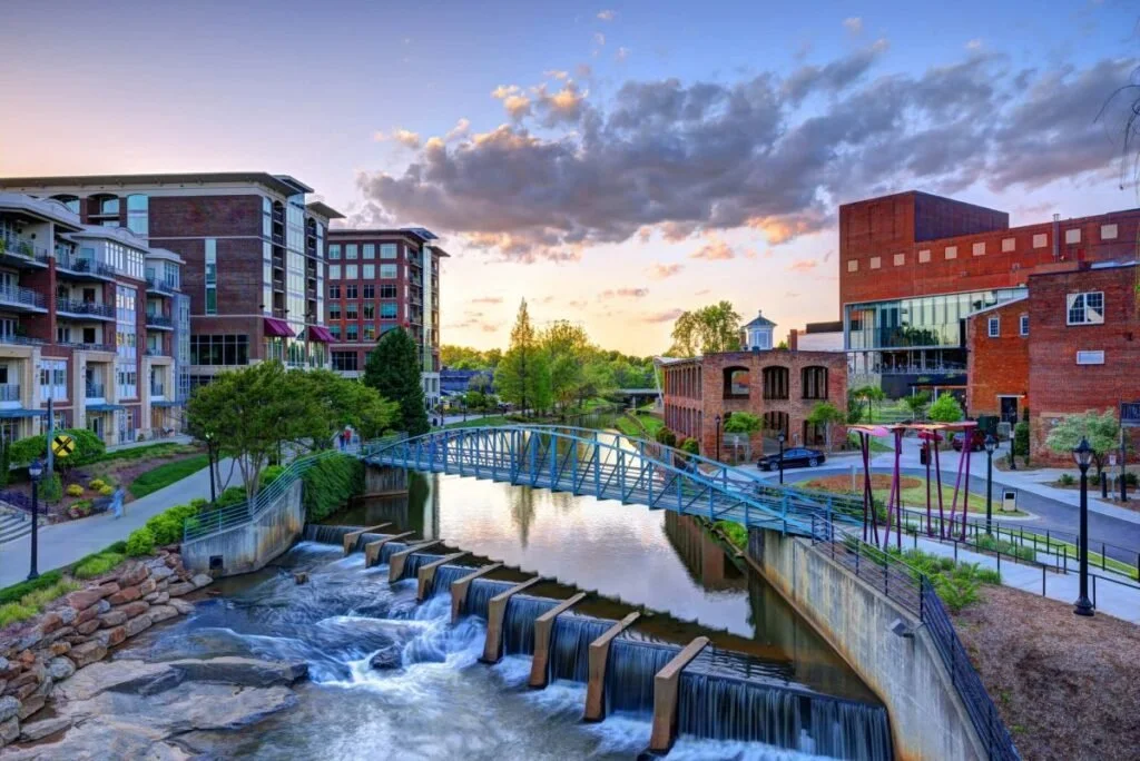 A cityscape with a river, a small waterfall, a blue pedestrian bridge, modern apartment buildings, trees, and a brick building under a partly cloudy sky at sunset.