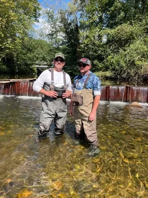 Two men fishing in a shallow river with a small waterfall in the background, surrounded by trees.