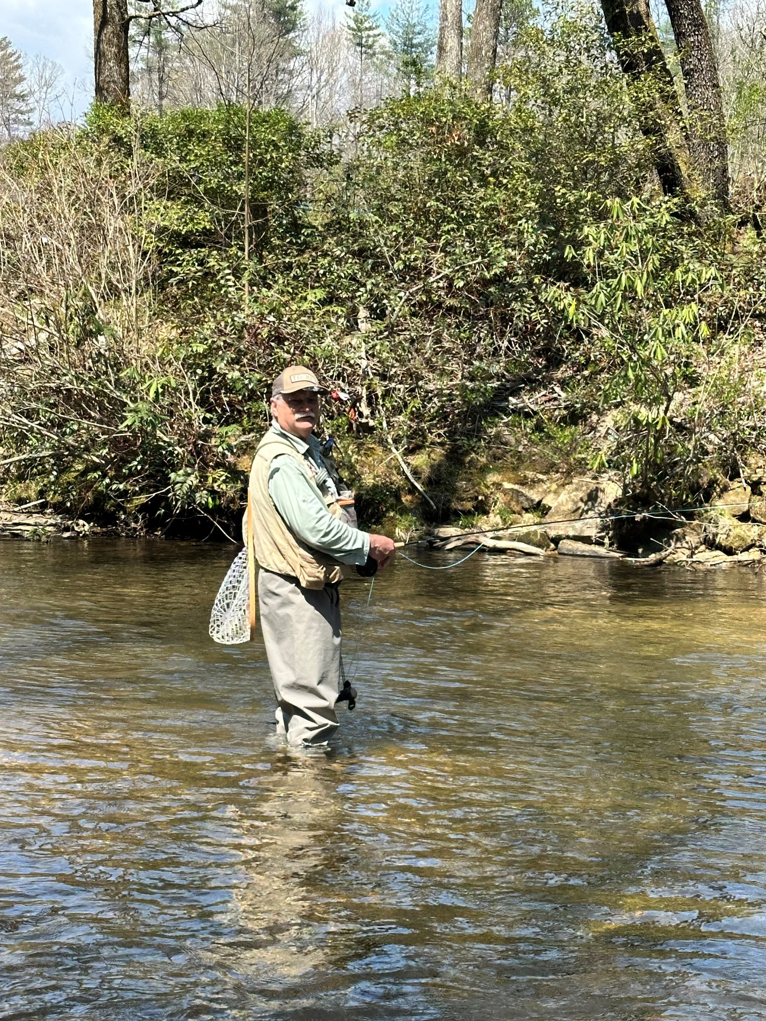 A man standing in a shallow river, holding a fishing pole, with dense bushes and trees in the background.