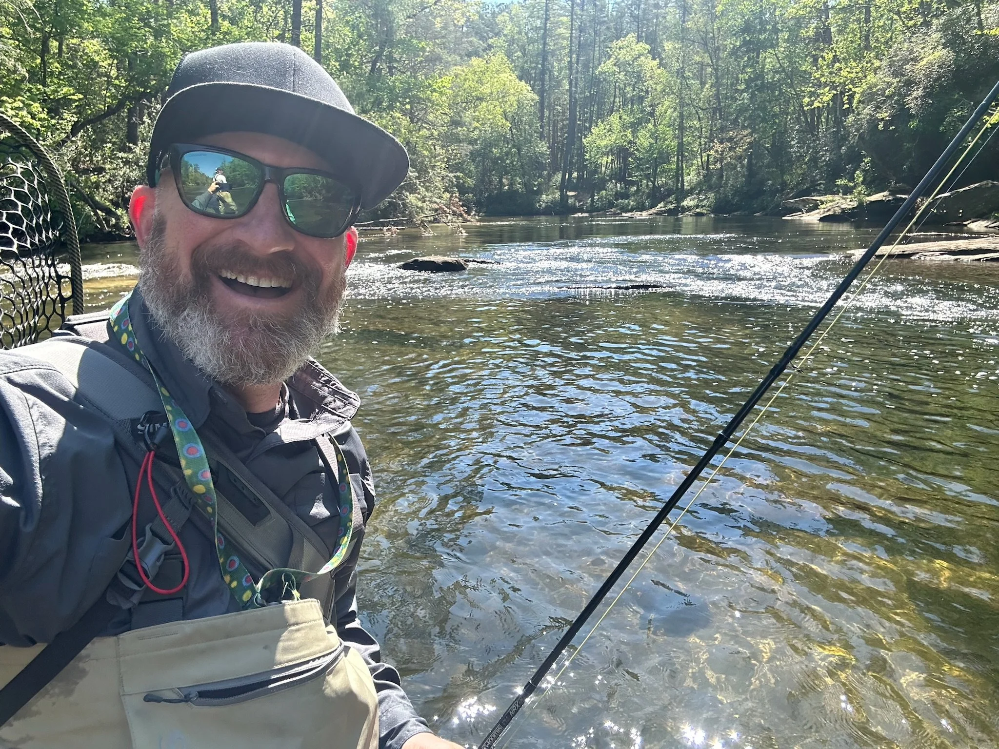 A man with a hat and sunglasses smiling while fishing in a river surrounded by lush green trees.