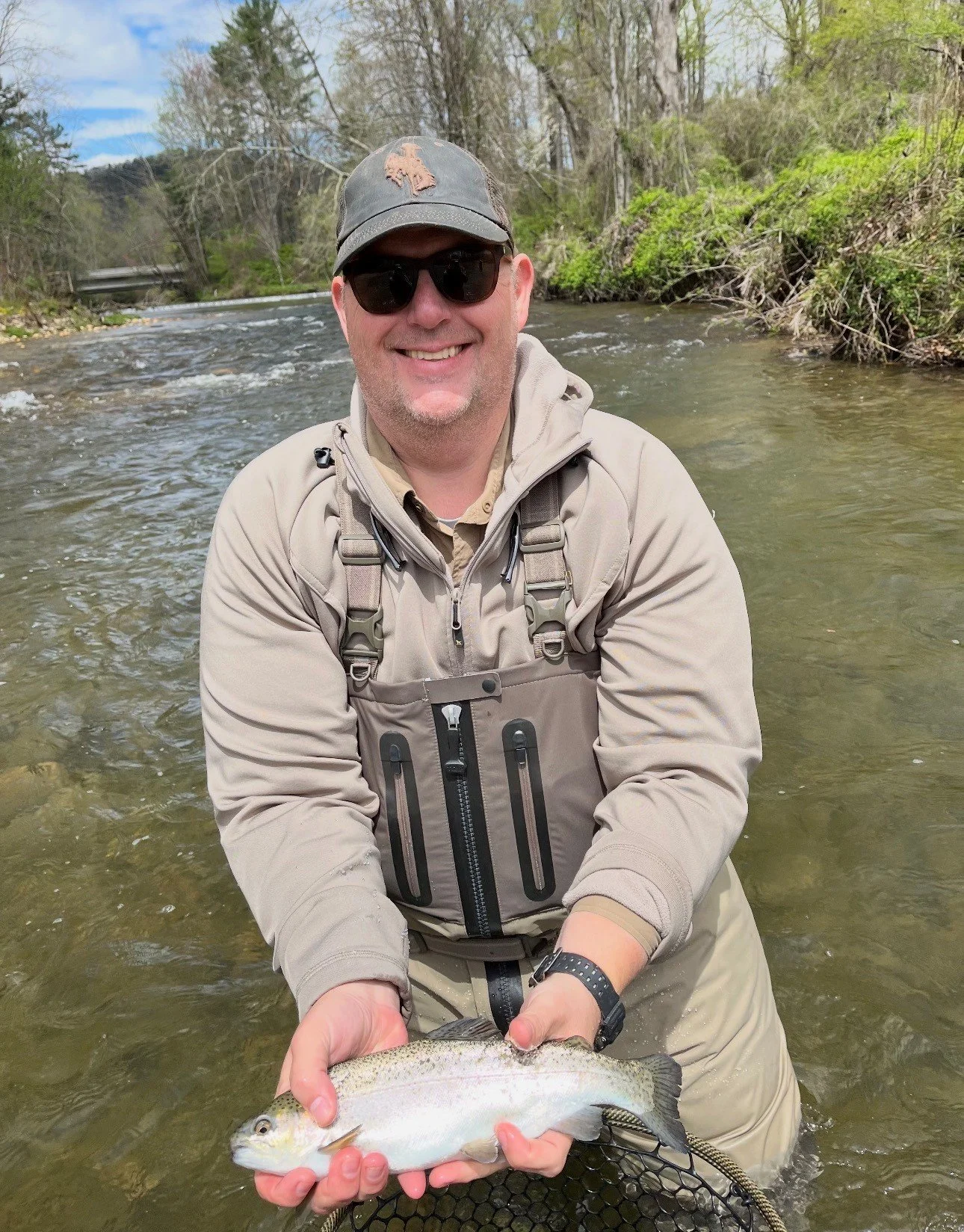 A man in fishing gear standing in a river, holding a fish he caught, smiling, with trees and a cloudy sky in the background.