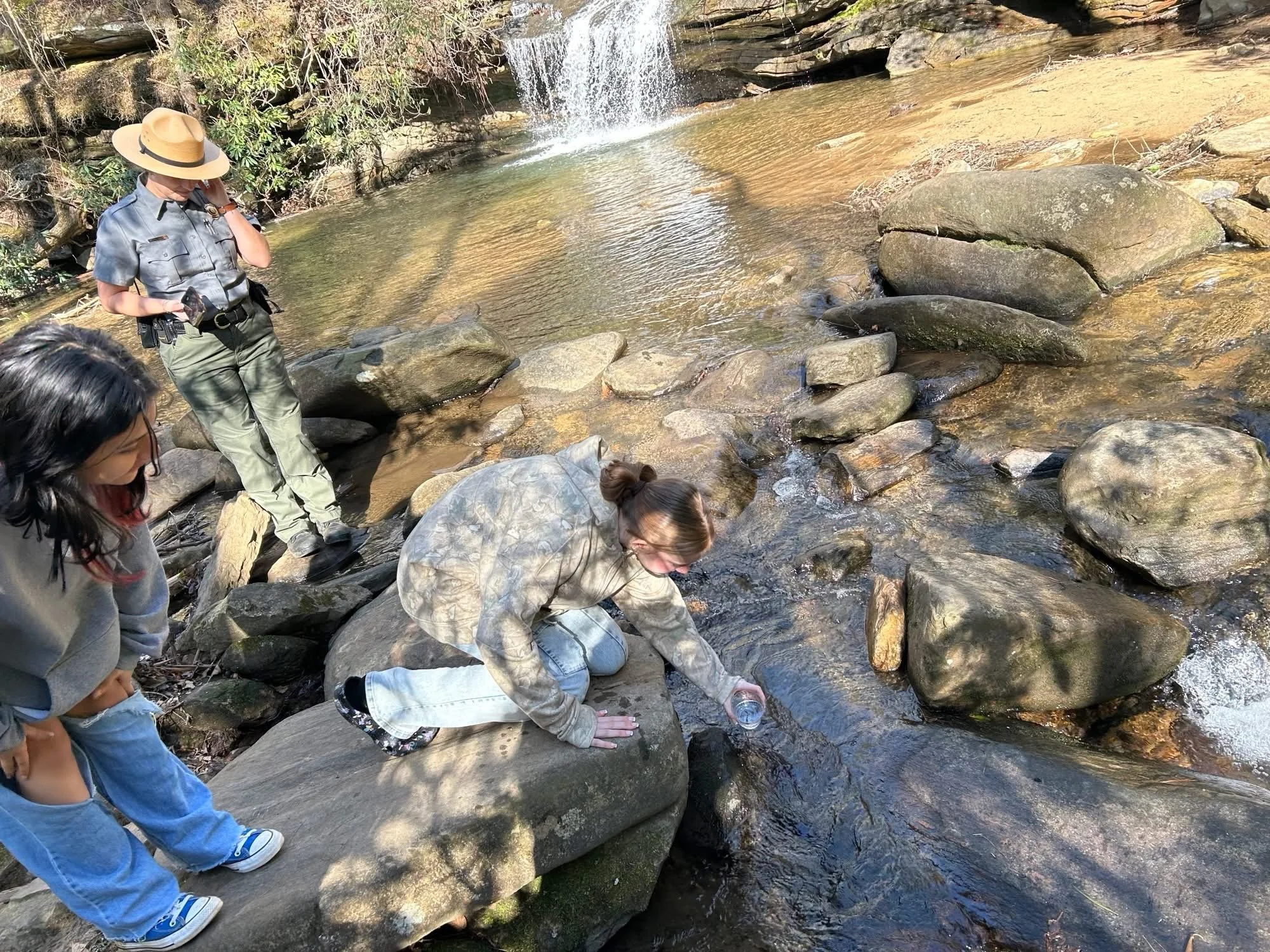 Children and a park ranger by a small waterfall in a rocky creek, surrounded by trees.