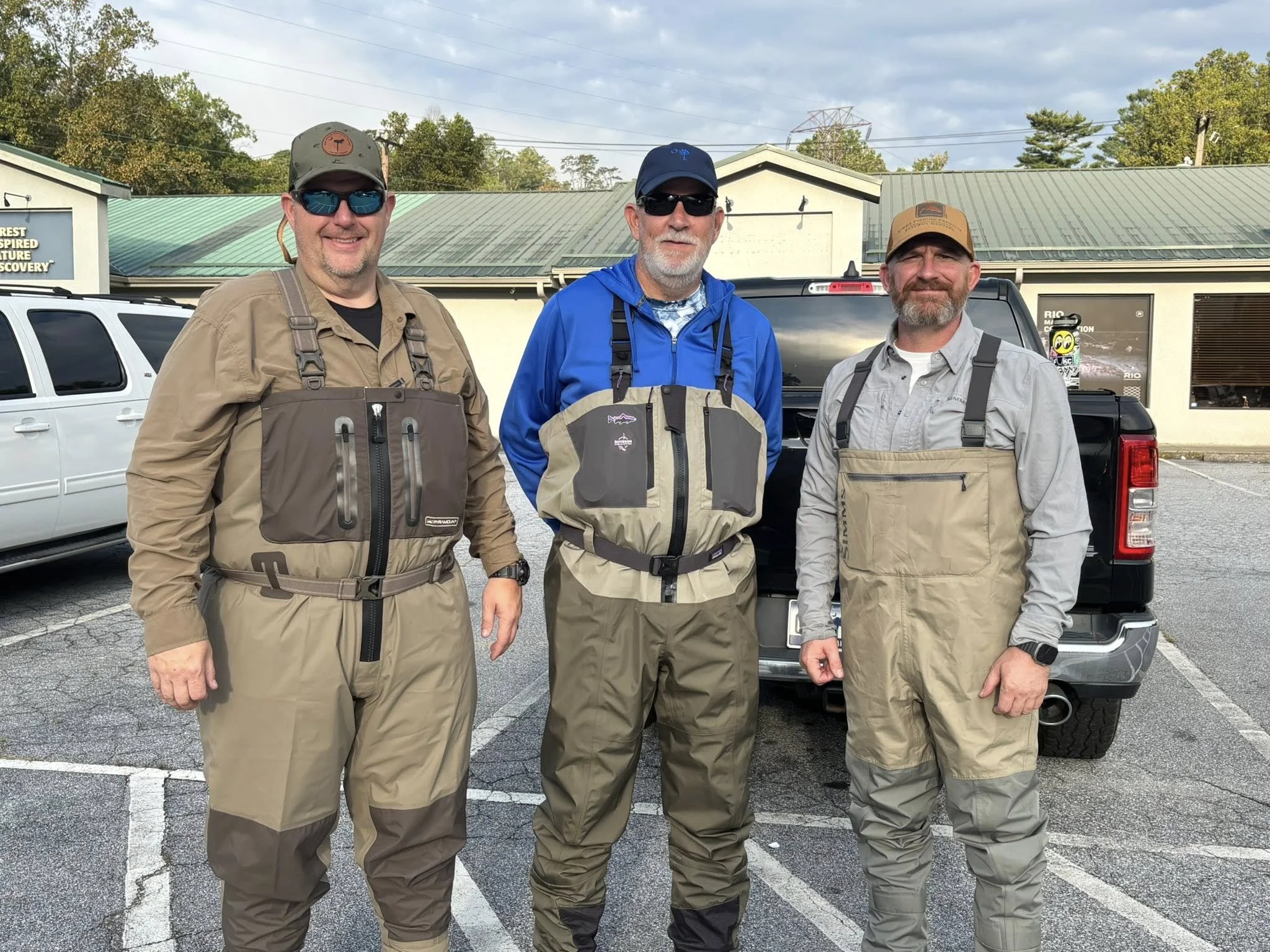 Three men dressed in fishing gear standing in a parking lot in front of a truck and other vehicles, with buildings and trees in the background.