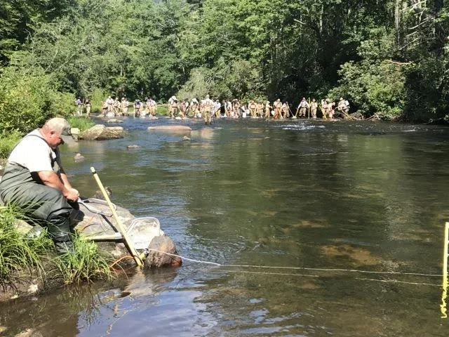 A man fishing in a river with many people standing along the other side of the riverbank in a wooded area.