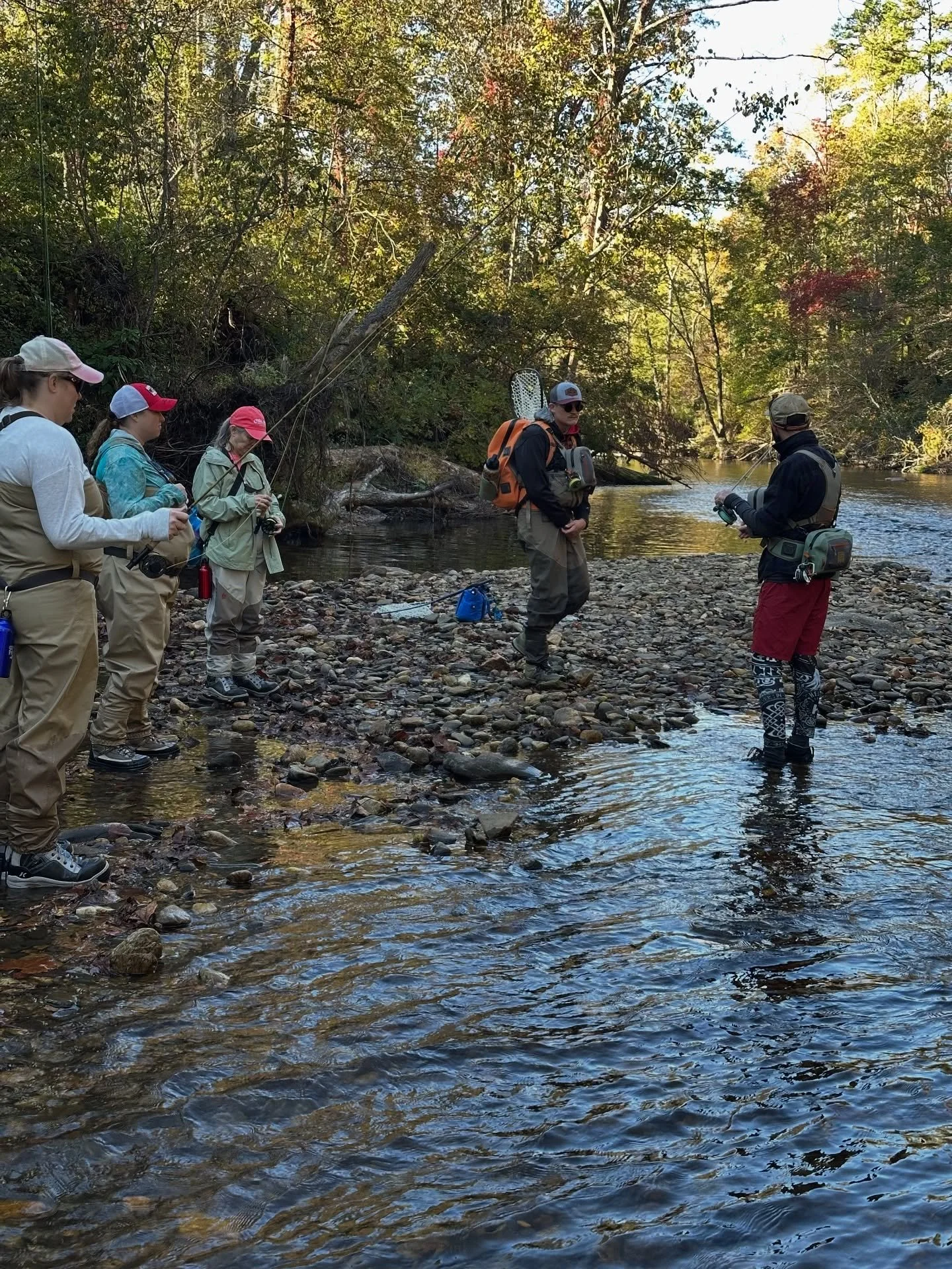 Group of people fishing in a shallow river surrounded by trees with fall foliage.