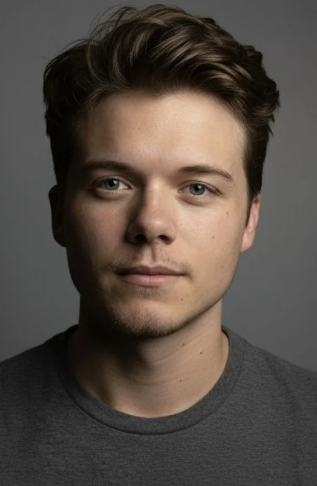 Close-up portrait of a young man with light skin, brown hair, and blue eyes, wearing a gray shirt, against a gray background.