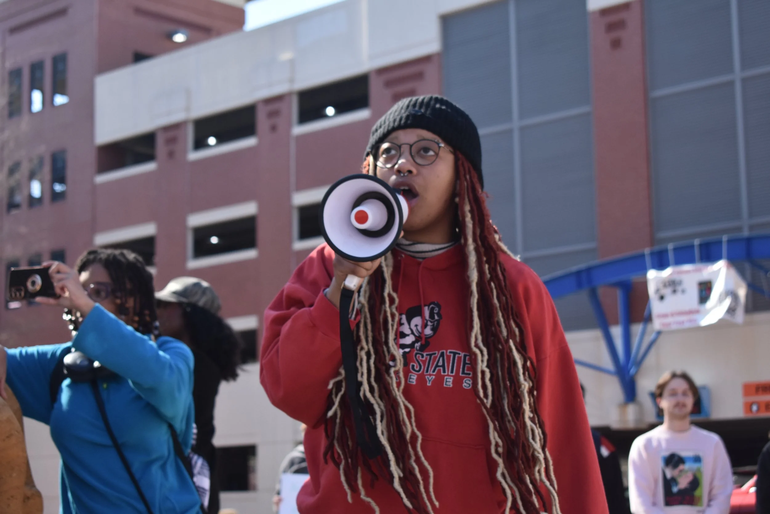 A person with long dreadlocks, glasses, and a black beanie, speaking into a megaphone during a protest or rally outside a modern building.