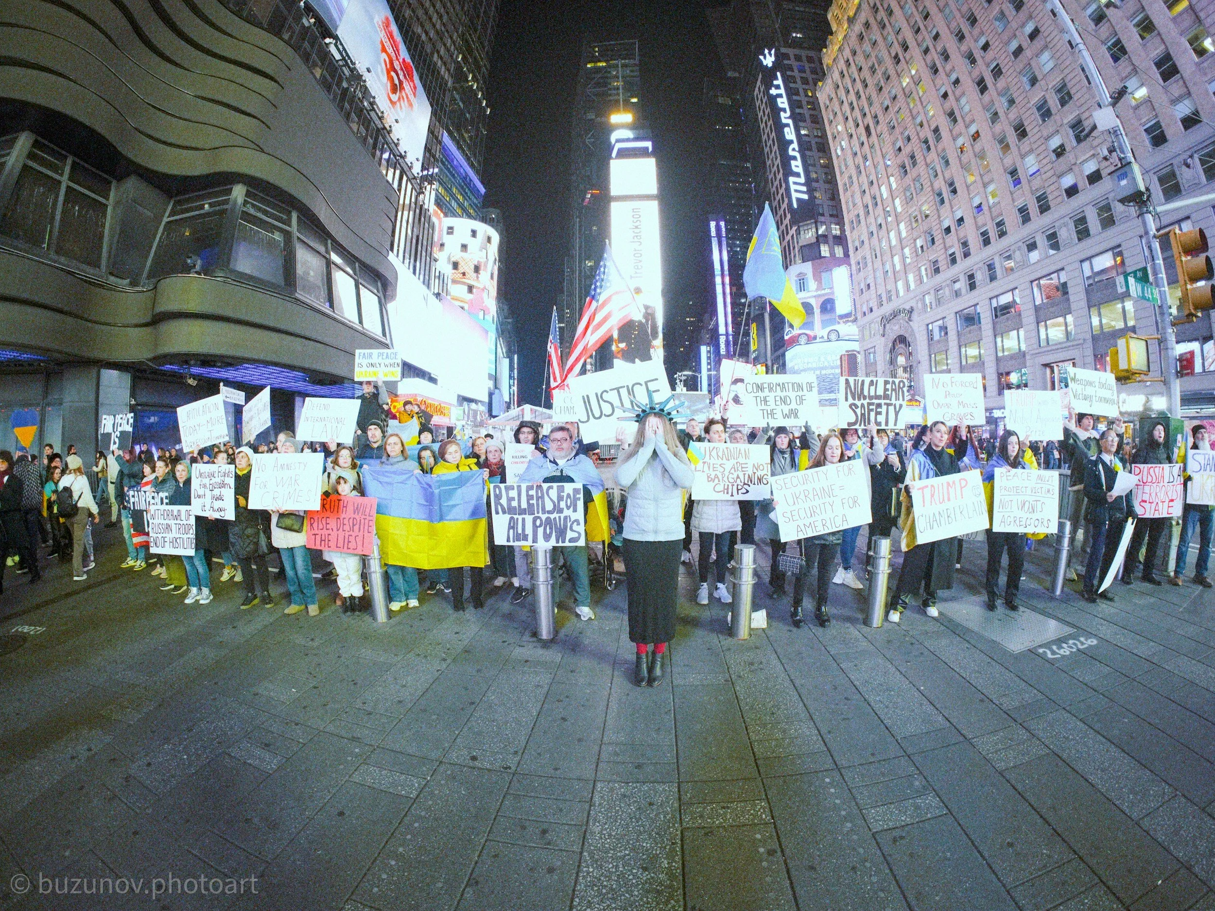 Protesters holding signs and flags in Times Square, New York City, at night. The protest involves various signs with messages about peace, justice, and Ukraine. The bright artificial city lights illuminate the scene, with tall buildings and billboards in the background.